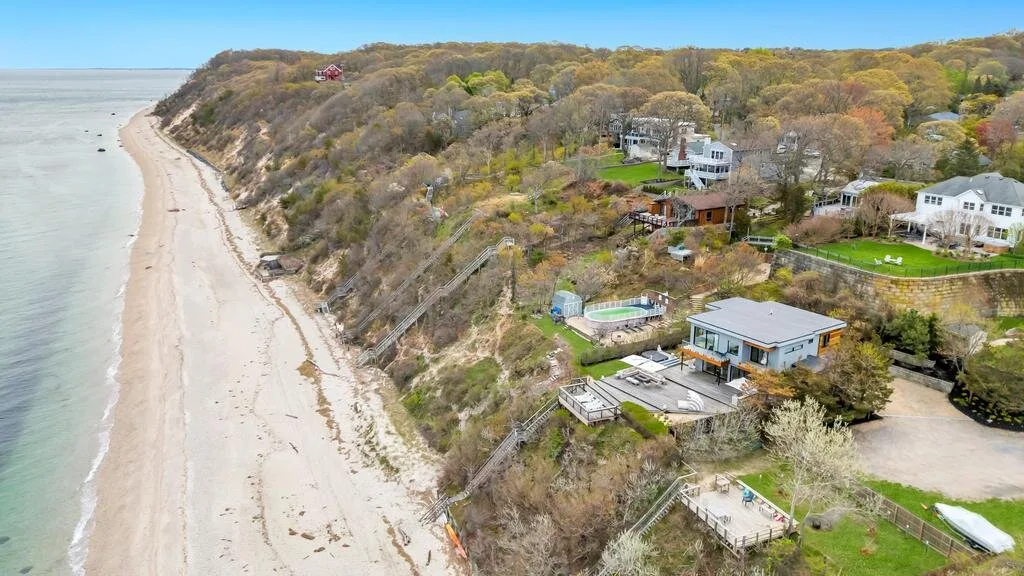 Vista aérea de una costa con playa de arena, acantilado arbolado y casas cercanas.