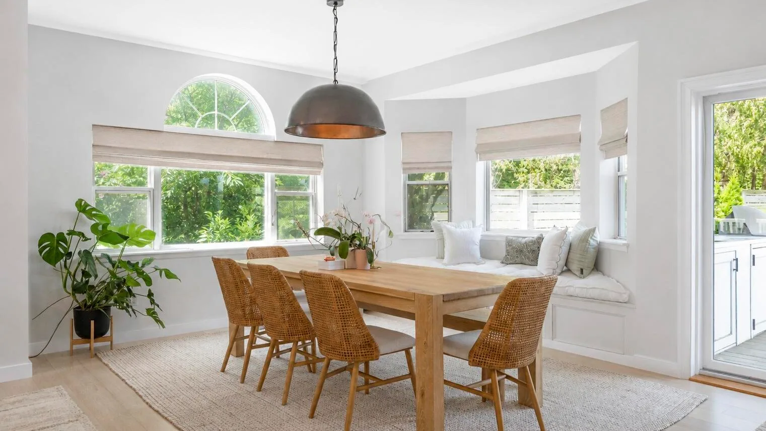Un comedor luminoso tiene una mesa de madera, asientos tejidos y grandes ventanas.