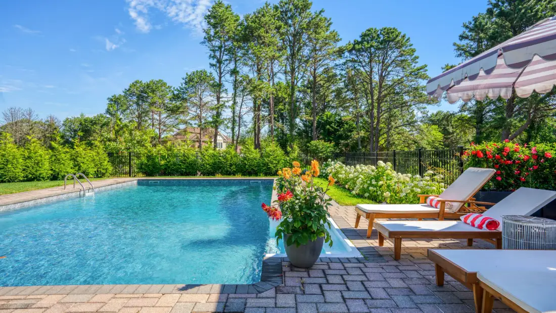 An outdoor swimming pool beside a paved patio with loungers and flowering plants.
