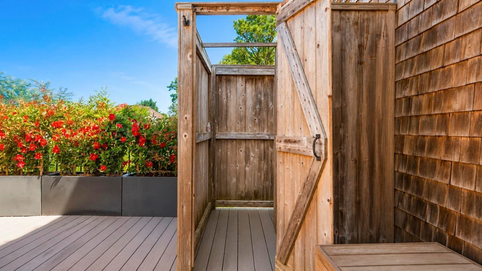 Una ducha exterior de madera se abre a una terraza junto a plantas con flores.
