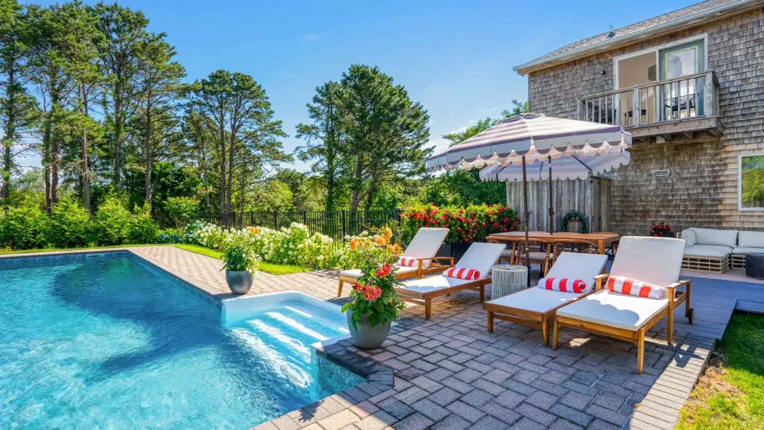 Una piscina exterior junto a un patio pavimentado con tumbonas y una sombrilla.