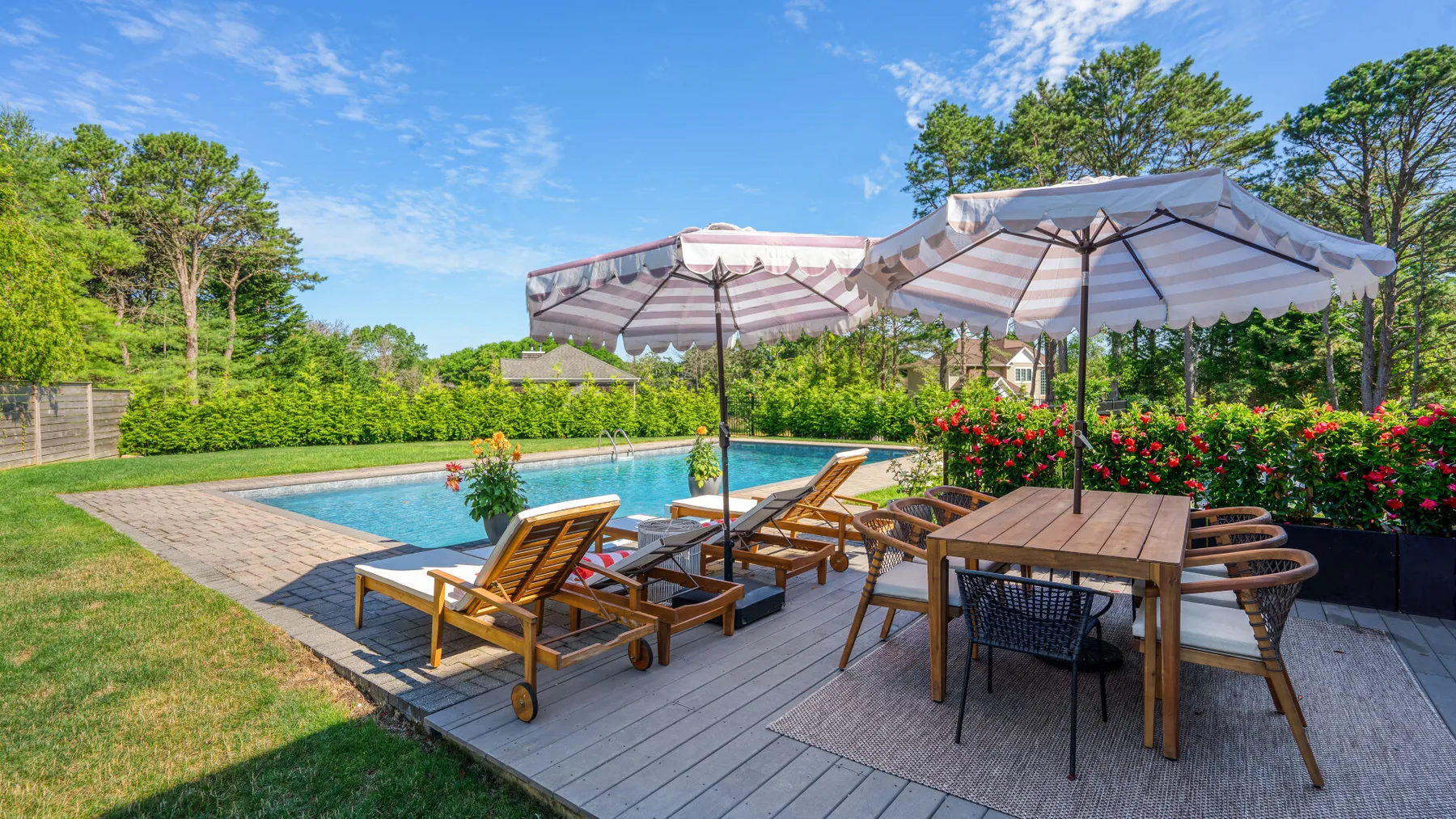 An outdoor poolside patio with wooden seating, umbrellas, and flowering plants.