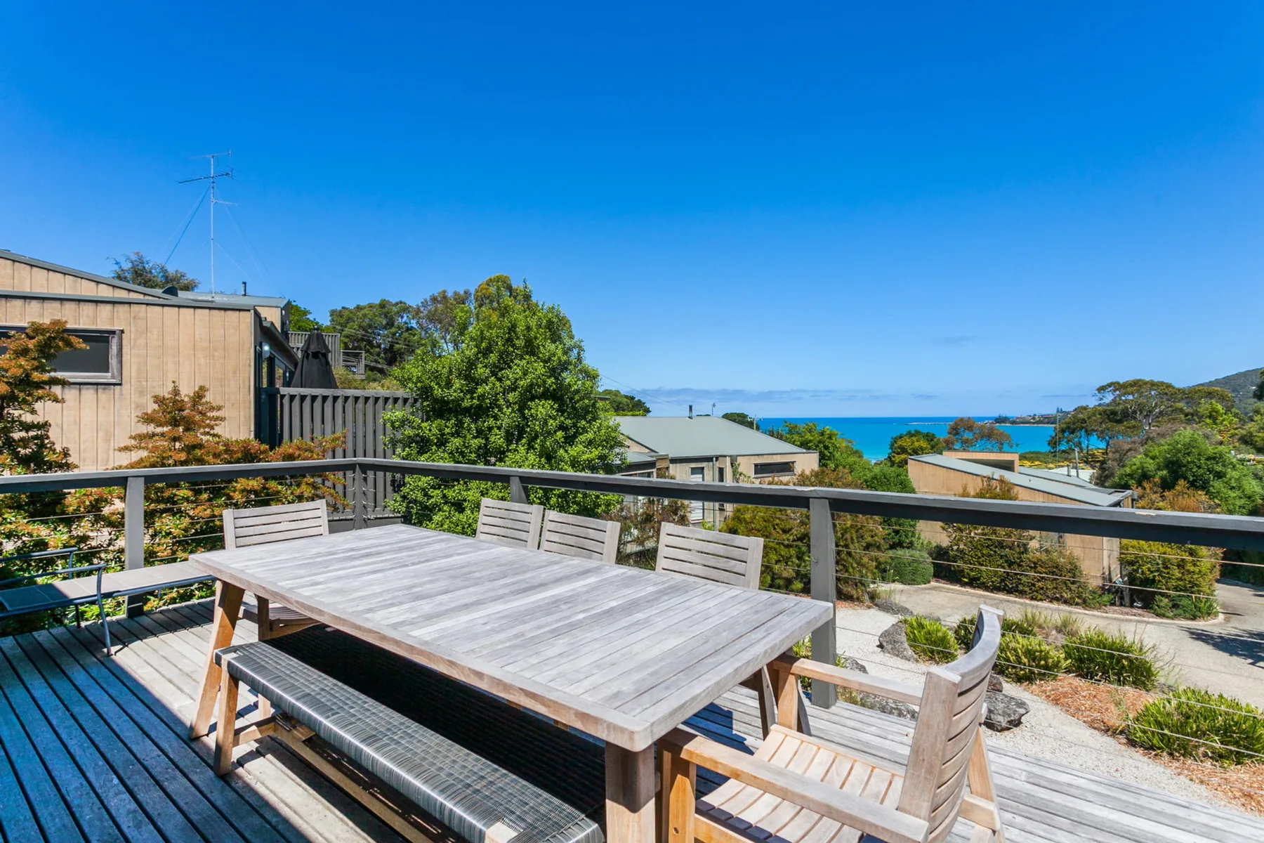 A timber outdoor deck has a dining table and seating overlooking the ocean.