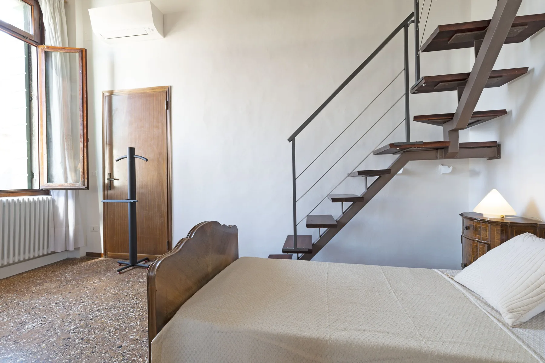 A bedroom with a bed and an open staircase beside the window.