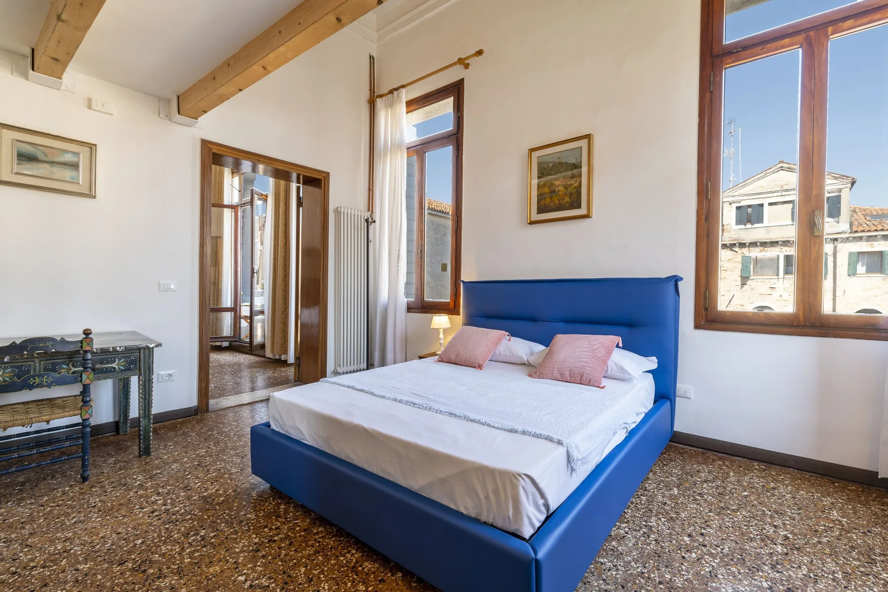 A bedroom with a blue bed, white bedding, and tall windows.