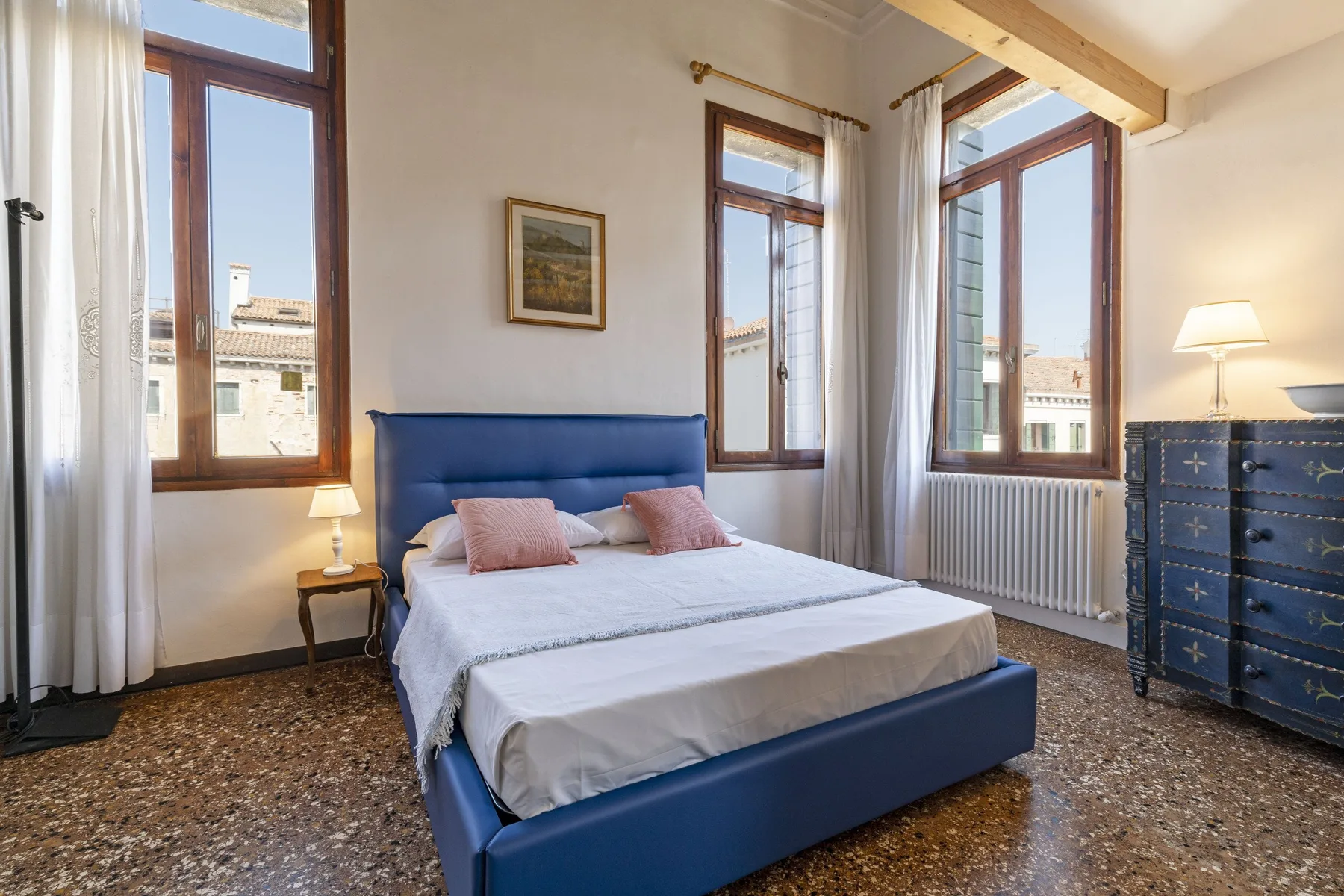 A bedroom with a blue bed, tall windows, and a dresser.