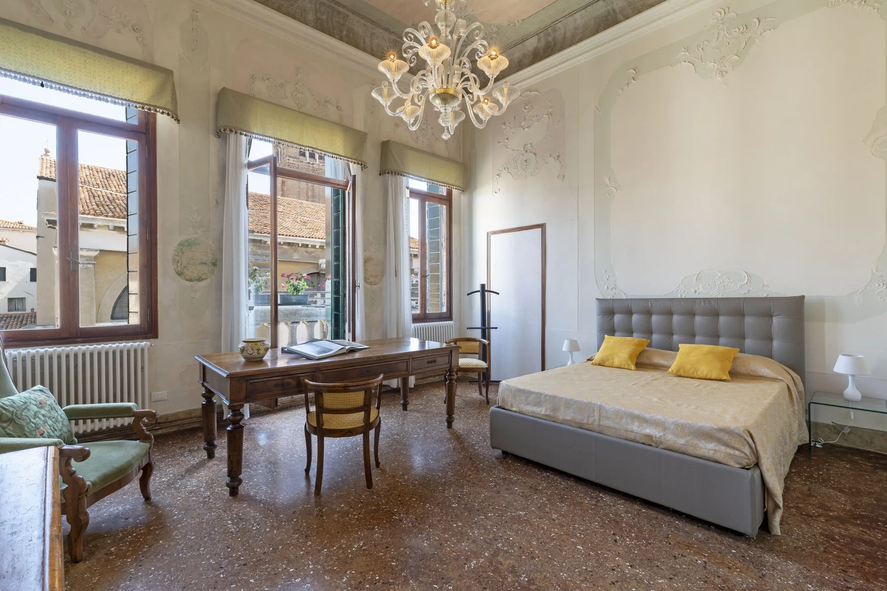 A bedroom with a bed, a desk, and tall windows overlooking nearby buildings.