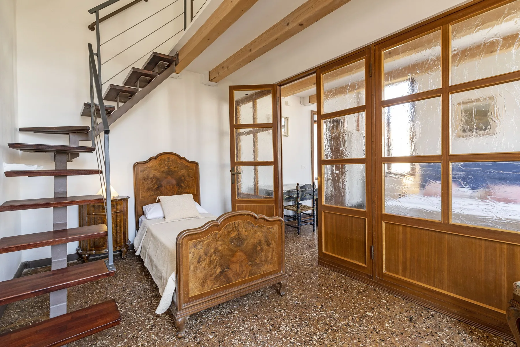 A bedroom with a single bed beneath an open staircase and wood-framed glass doors.