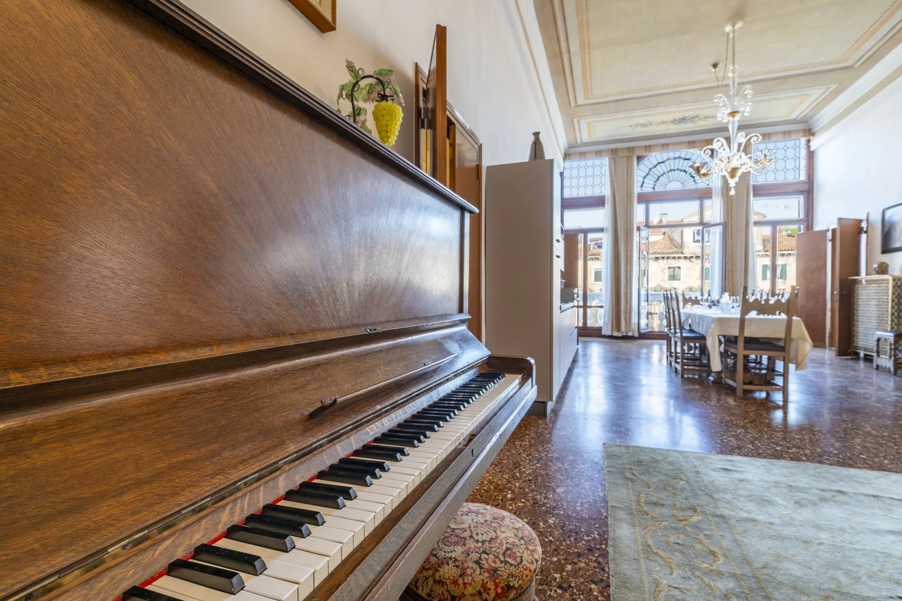 A piano stands in a bright dining room with tall windows and a table.