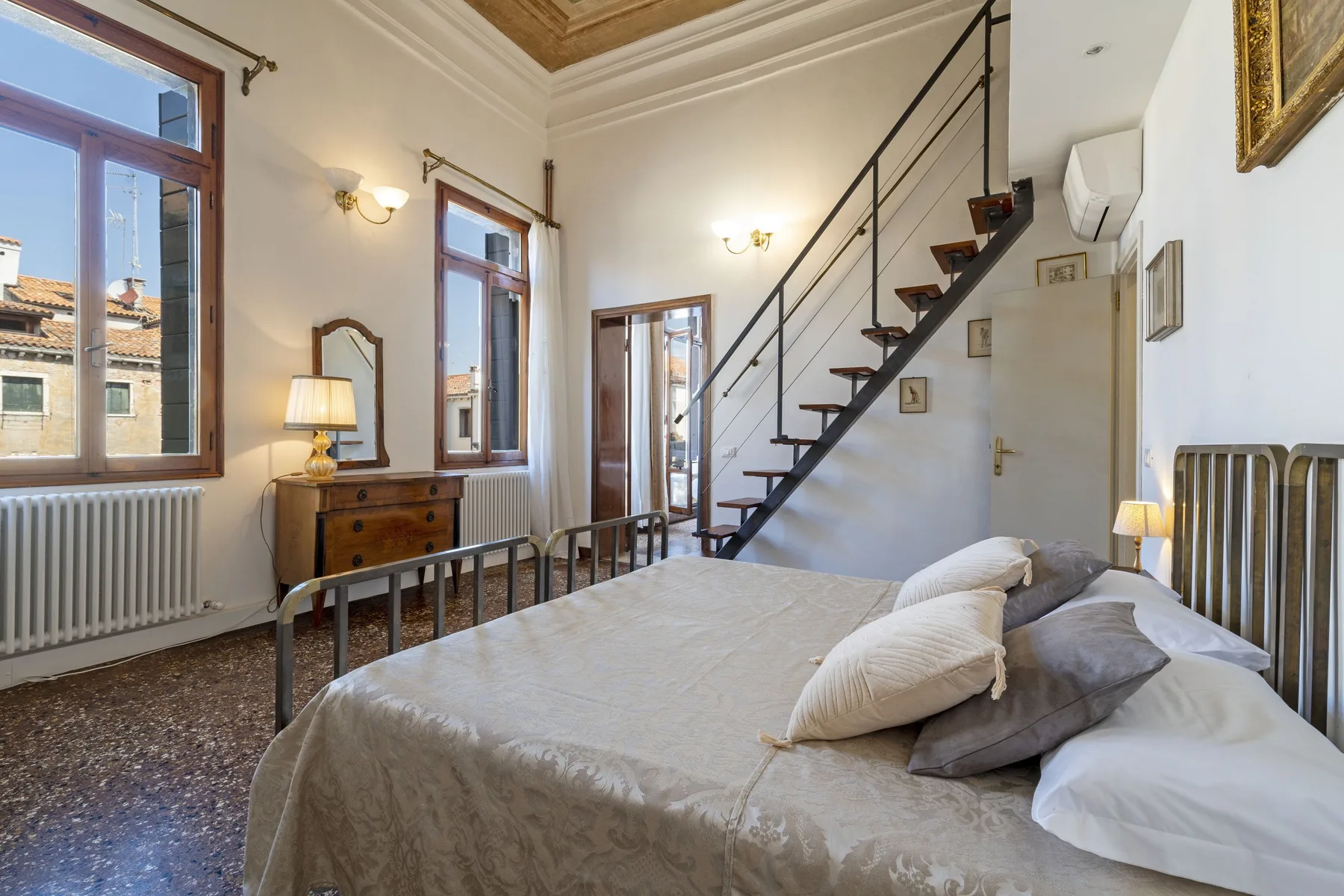 A bedroom with a large bed beneath a staircase and tall windows.