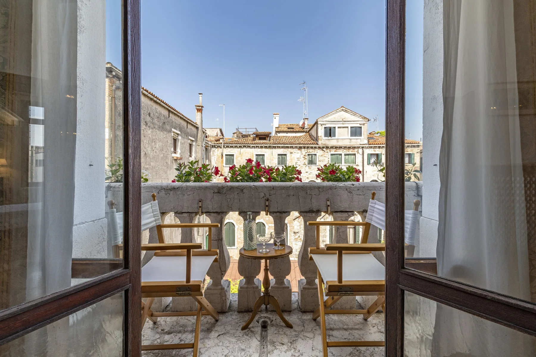 An open balcony with two seats and flowers overlooks neighboring buildings.
