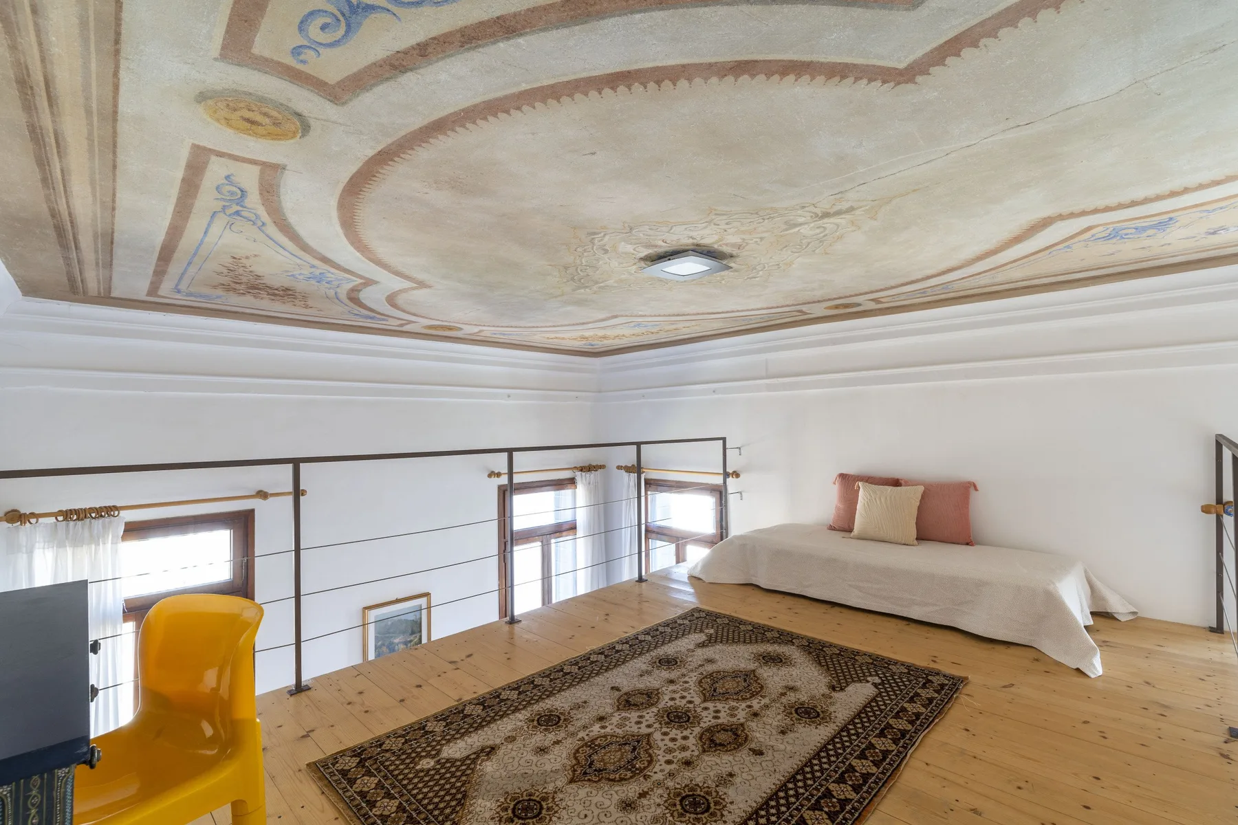 A bright attic room with a bed, patterned rug, and ornate painted ceiling.