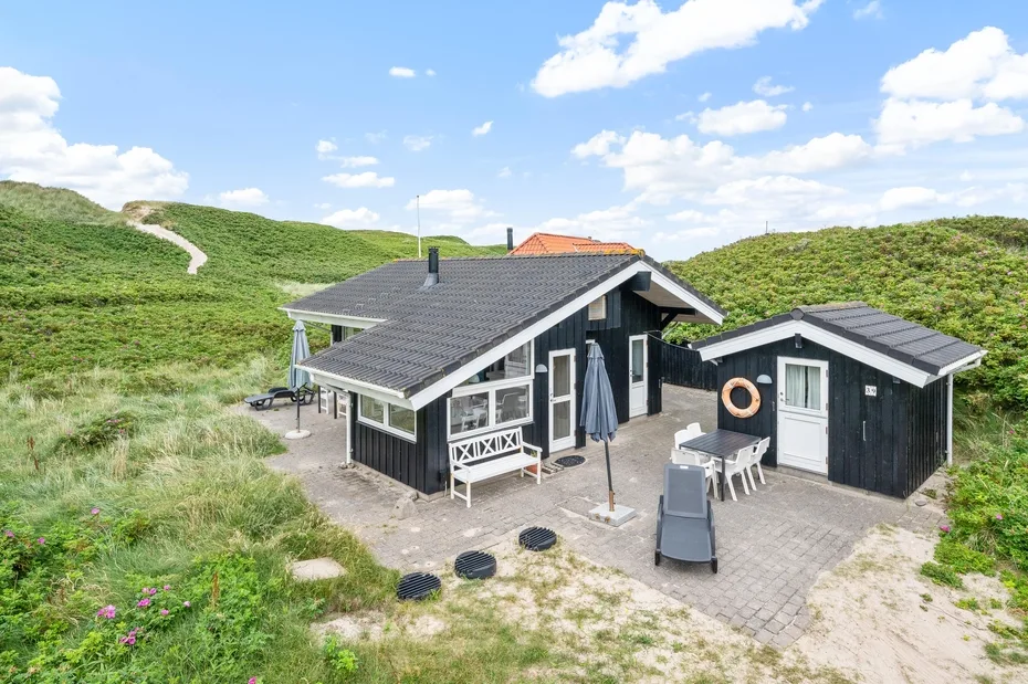 Una casa oscura se encuentra entre dunas verdes bajo un cielo despejado con nubes.