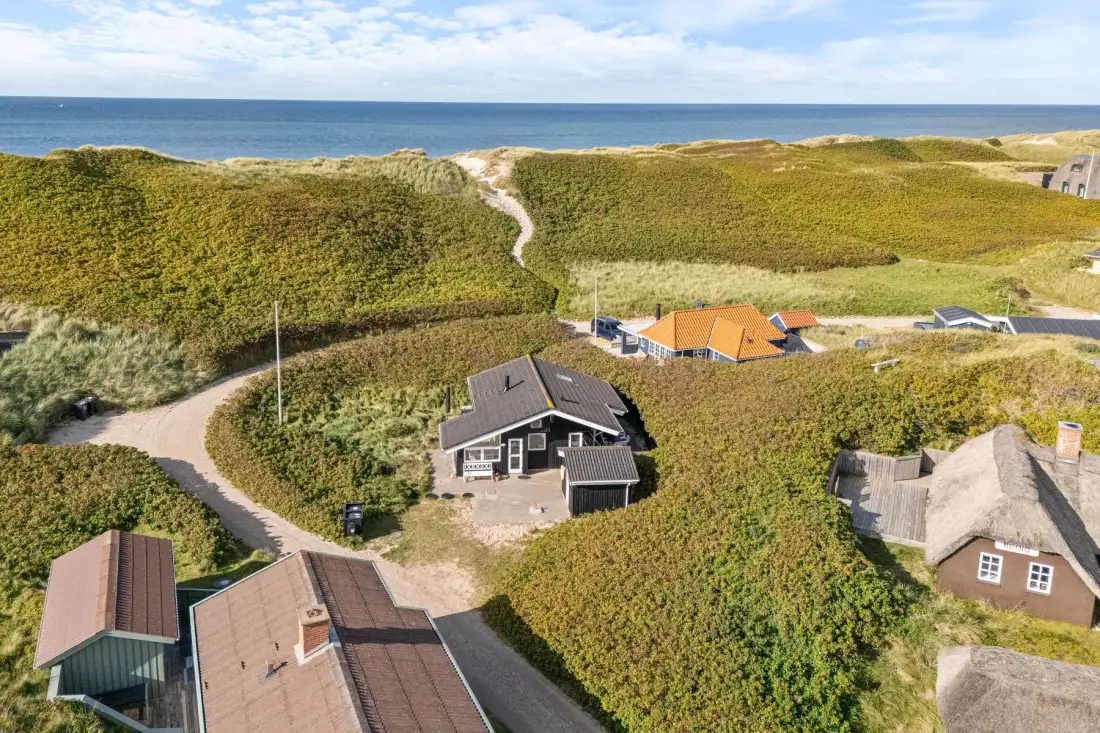 Vista aérea de casas vacacionales entre dunas cubiertas de hierba, con el mar al fondo.