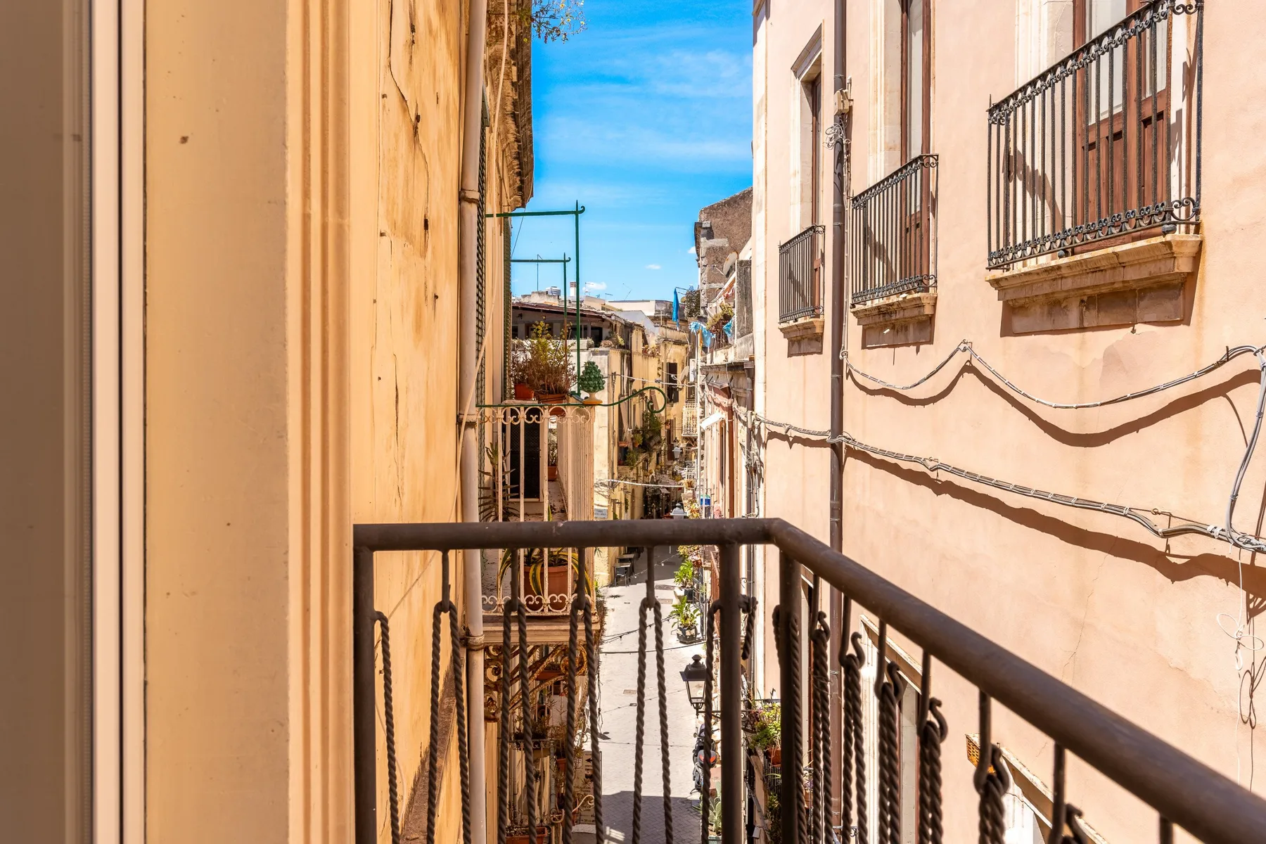 Una calle urbana estrecha vista desde un balcón, con edificios claros y barandillas de hierro.