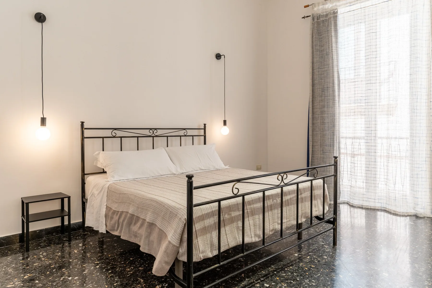Un dormitorio con una cama de metal, una mesita y cortinas junto a una ventana luminosa.