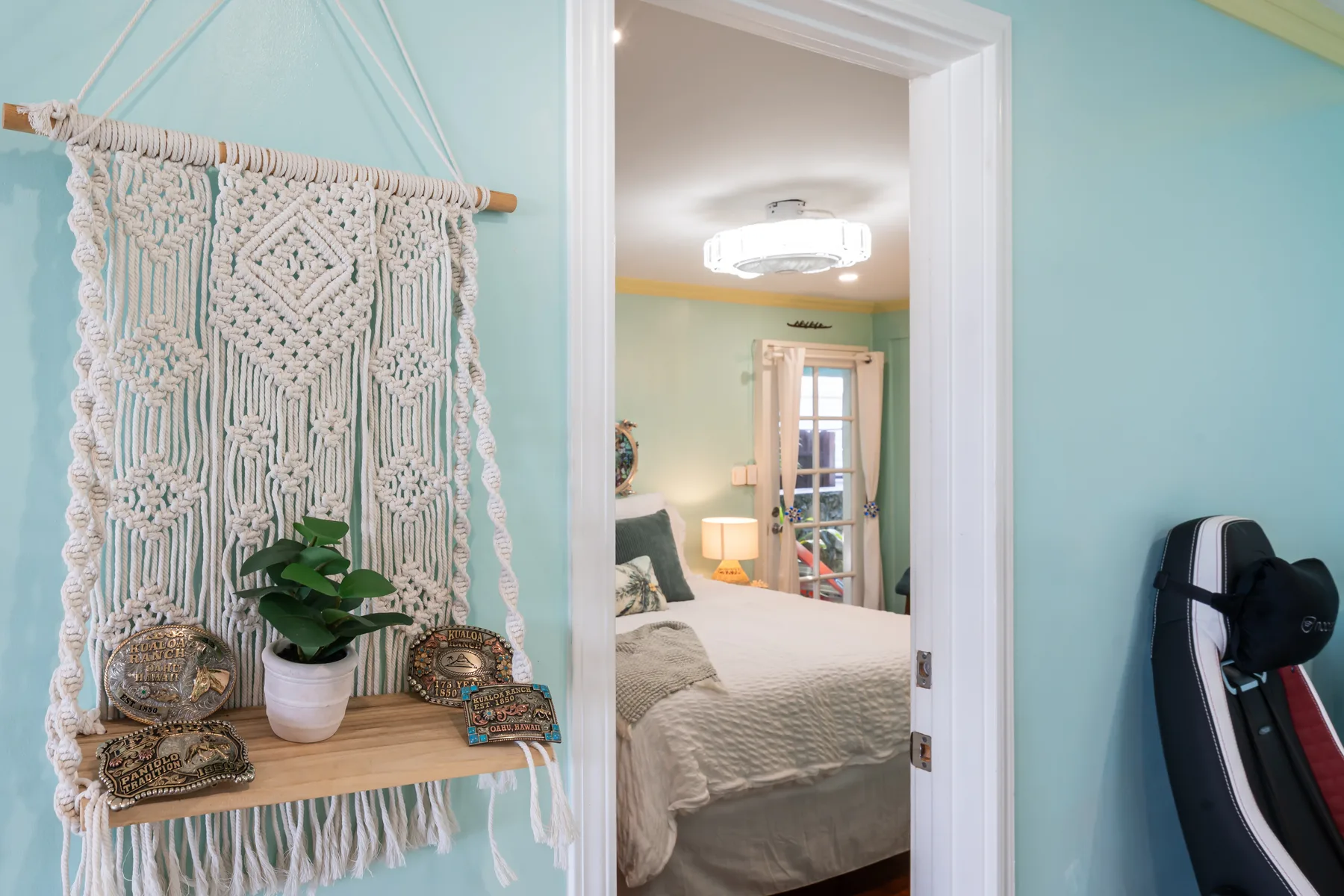 Se ve un dormitorio a través de una puerta, junto a un tapiz de macramé y una planta en maceta.