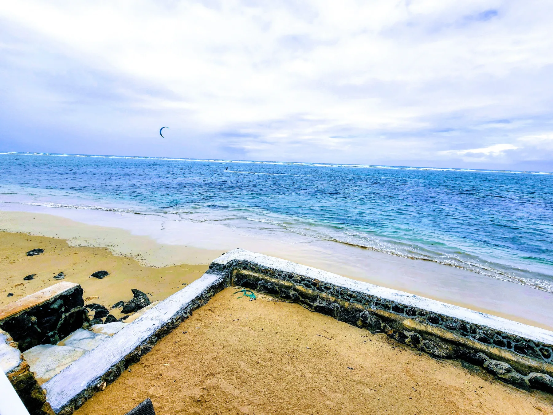 Una playa de arena junto al agua azul, con un muro bajo de piedra en primer plano.
