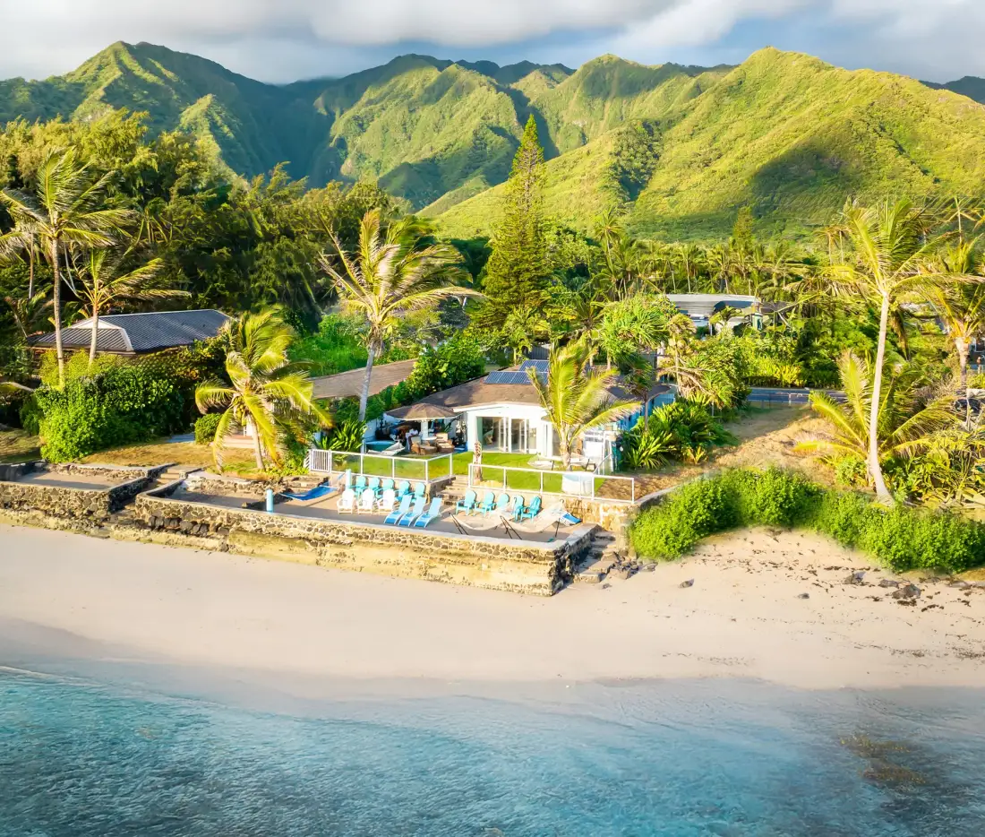 Un complejo frente a la playa con piscina y palmeras se encuentra bajo montañas verdes.