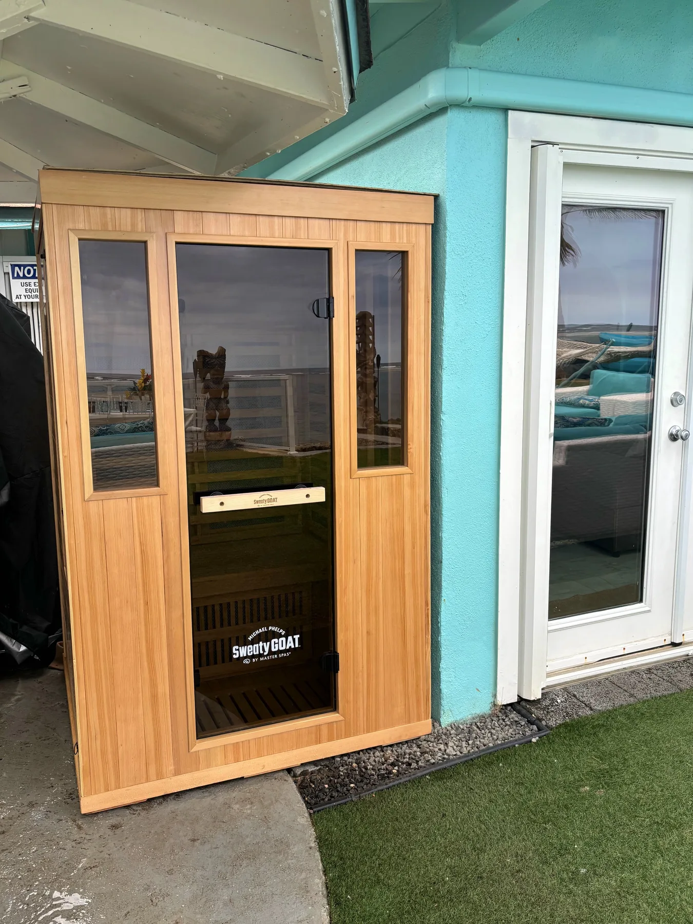 Una sauna de madera está junto a una puerta de vidrio en una pared exterior turquesa.