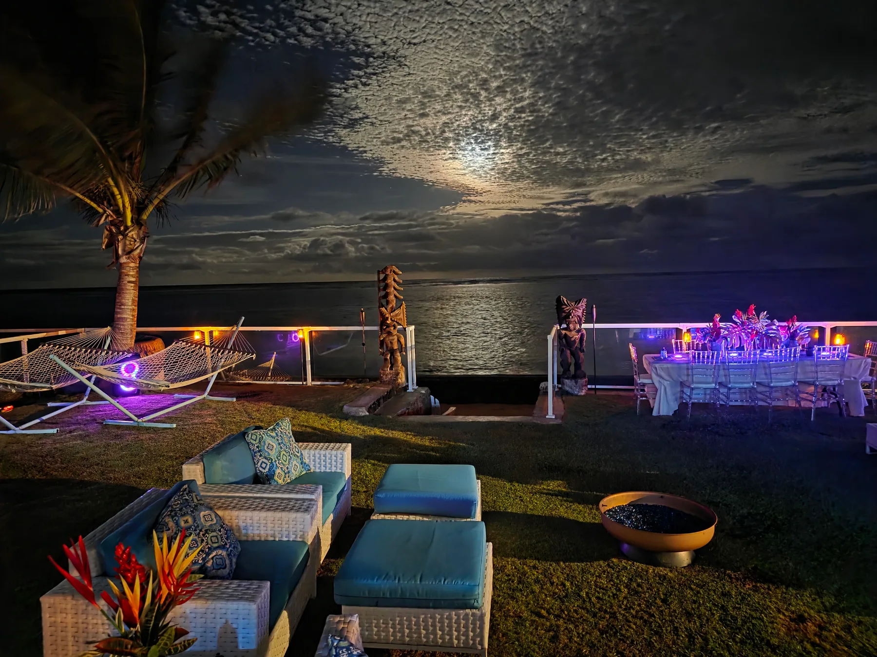 Una zona de estar al aire libre junto al agua, de noche, con asientos acolchados y una mesa iluminada.