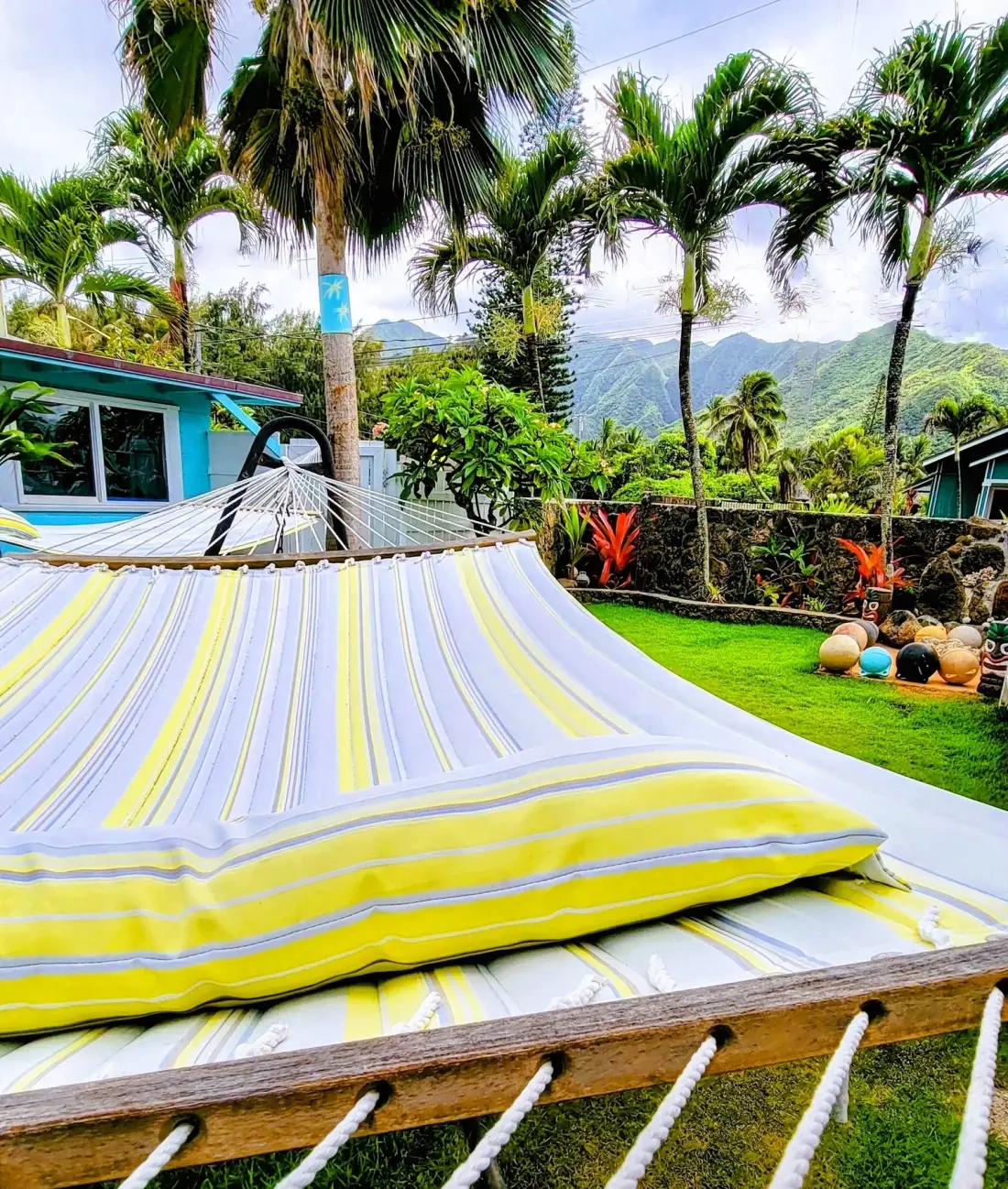 Una hamaca a rayas descansa en un jardín tropical con palmeras y montañas verdes.