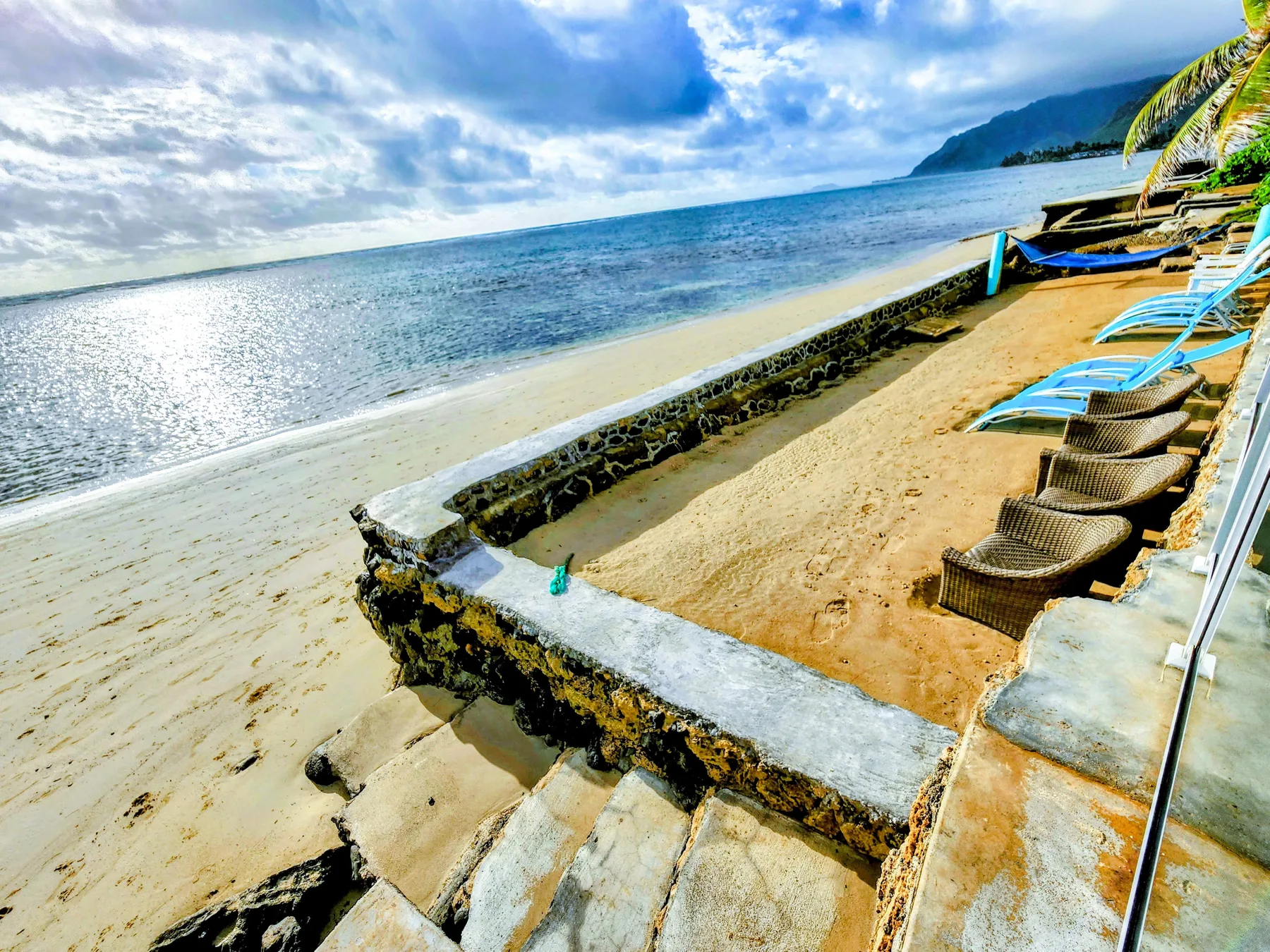 Una playa de arena con asientos junto a un muro bajo de piedra y aguas tranquilas.