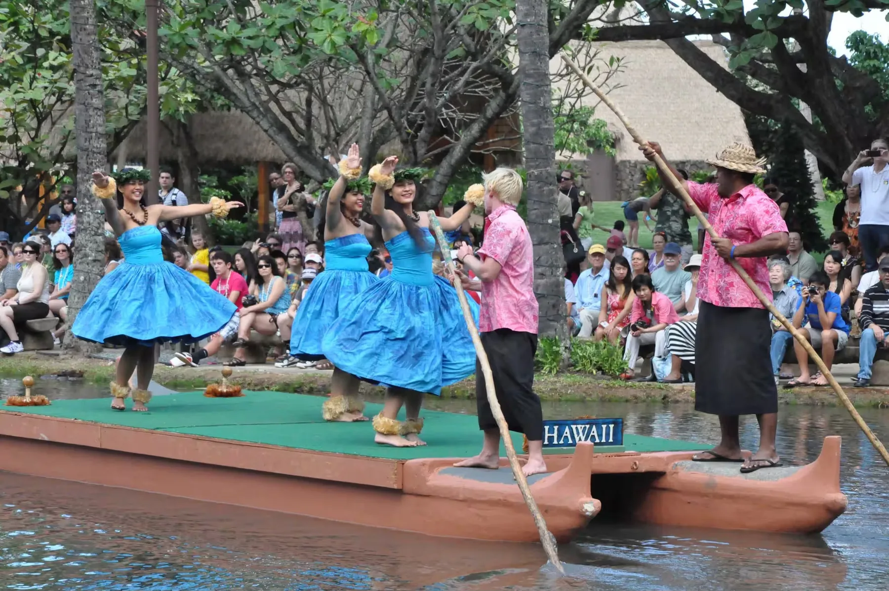 Artistas bailan en una plataforma junto al agua mientras el público observa desde la orilla.