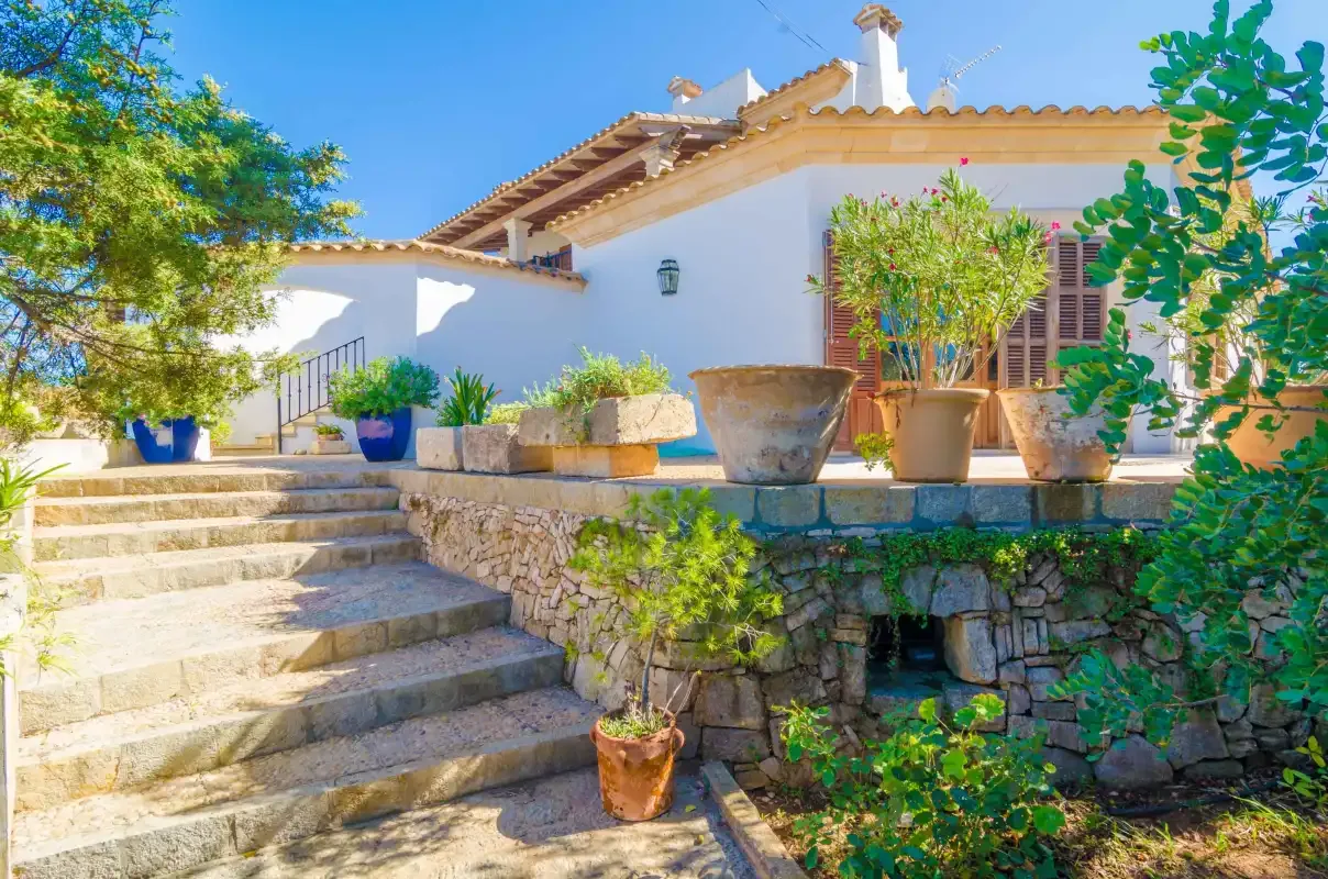 A white house with tiled roofs stands above stone steps and a planted terrace.