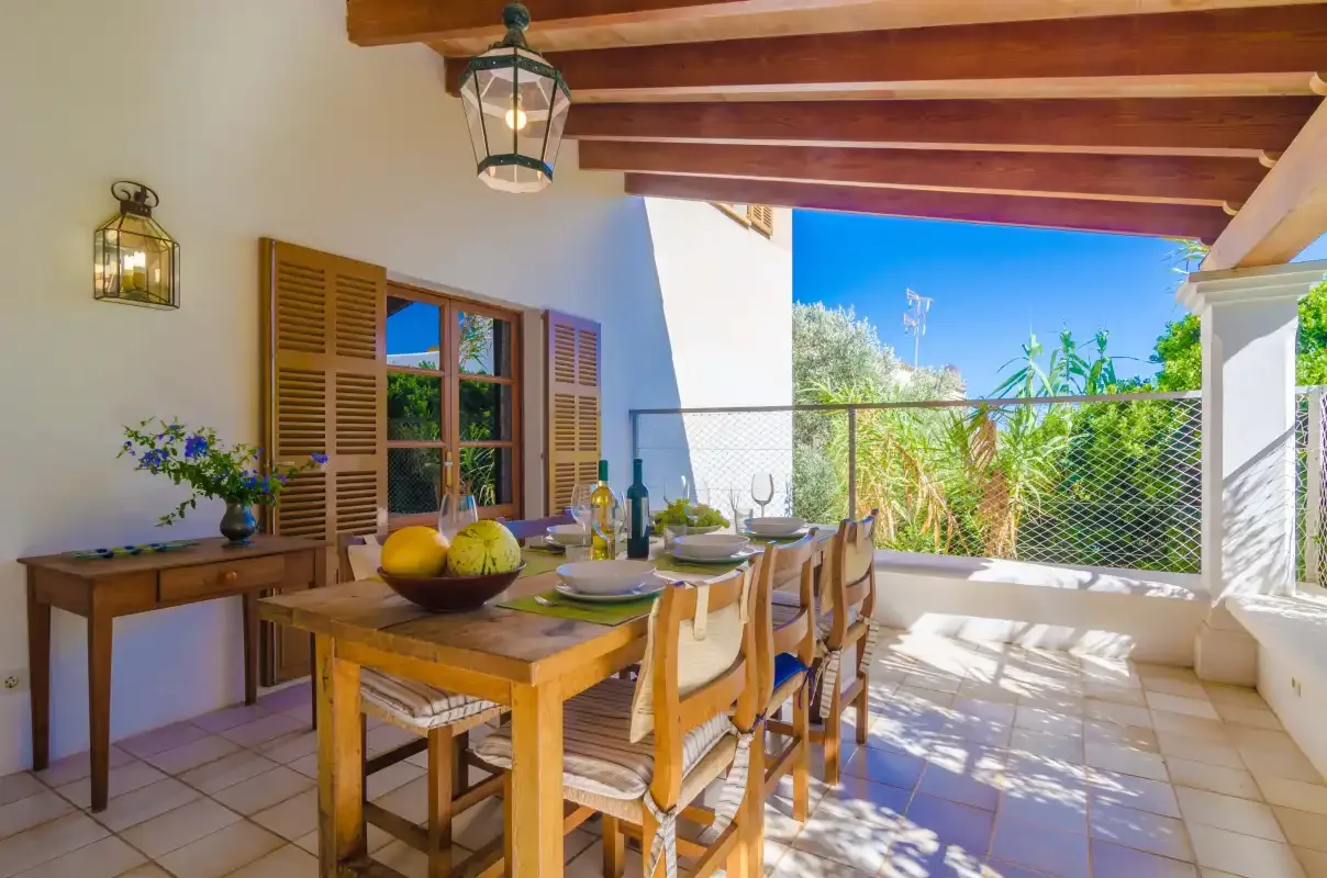 A covered outdoor dining area with a wooden table beside open shutters.