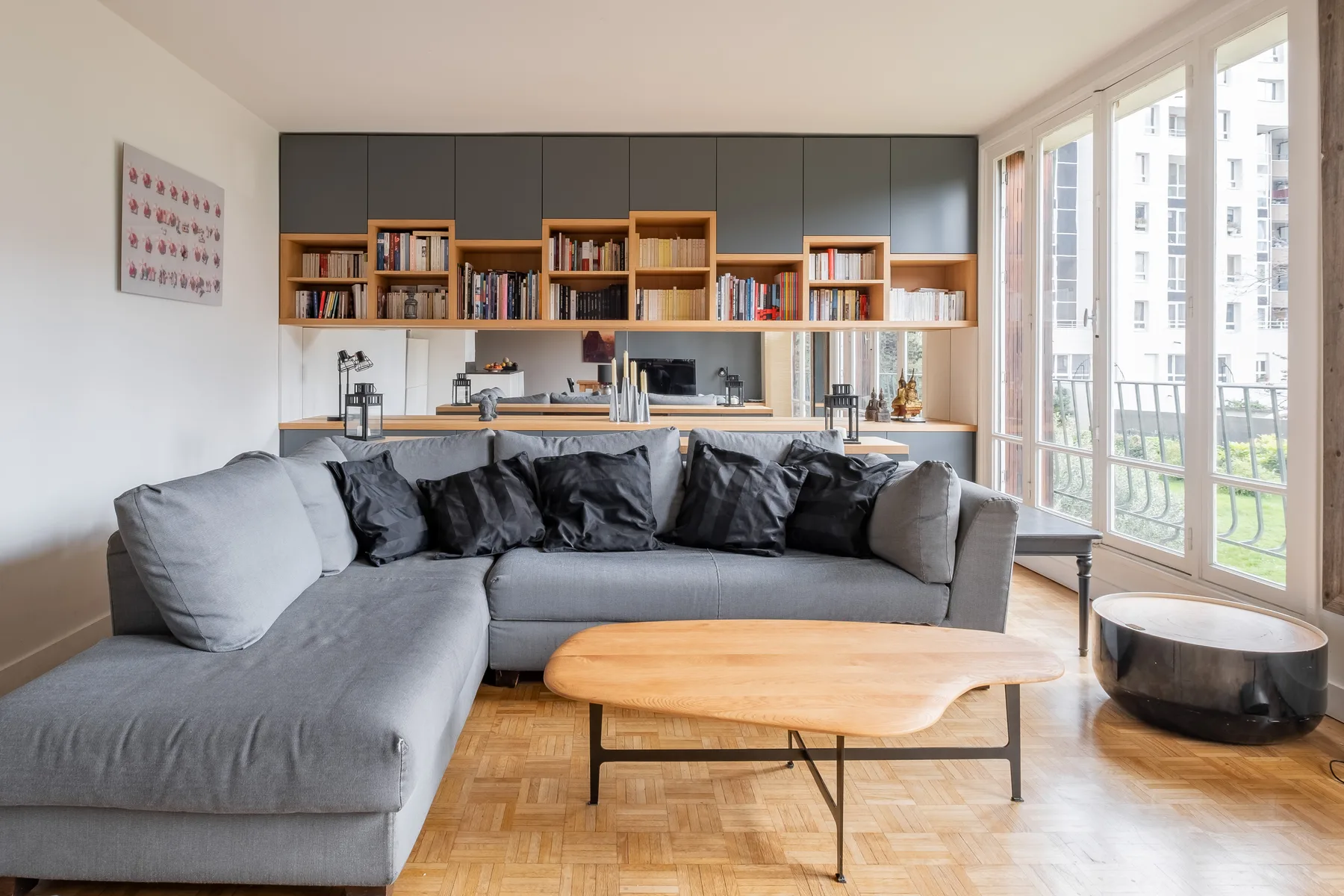 Una sala luminosa tiene un sofá seccional gris, una mesa de centro de madera y ventanales.