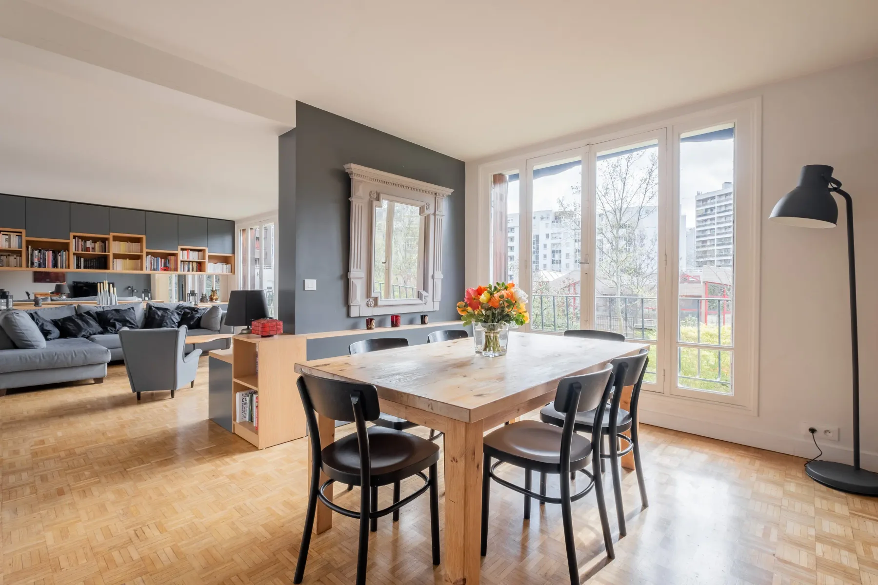 Comedor con una mesa de madera junto a grandes ventanas, abierto a una zona de estar.