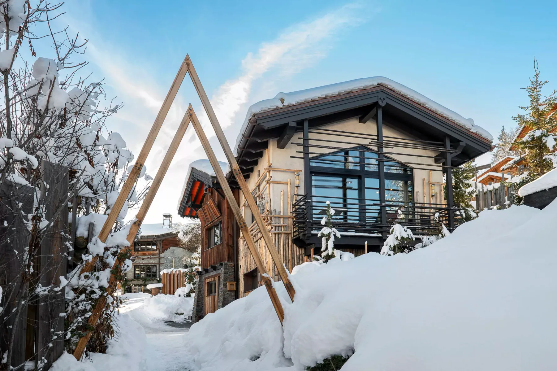 A modern chalet stands beside a snow-covered path, with trees and deep snow around it.