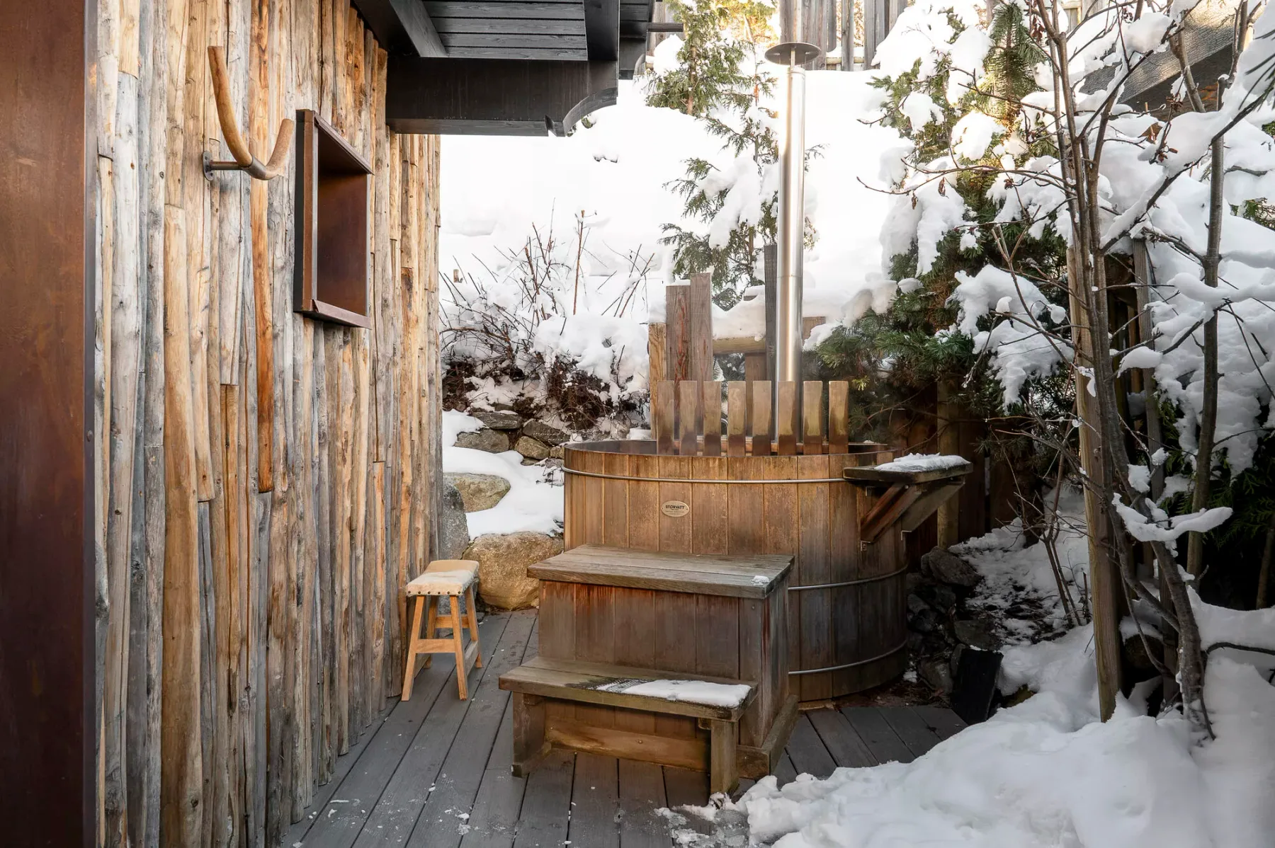 A wooden outdoor hot tub sits on a deck surrounded by snow-covered trees.