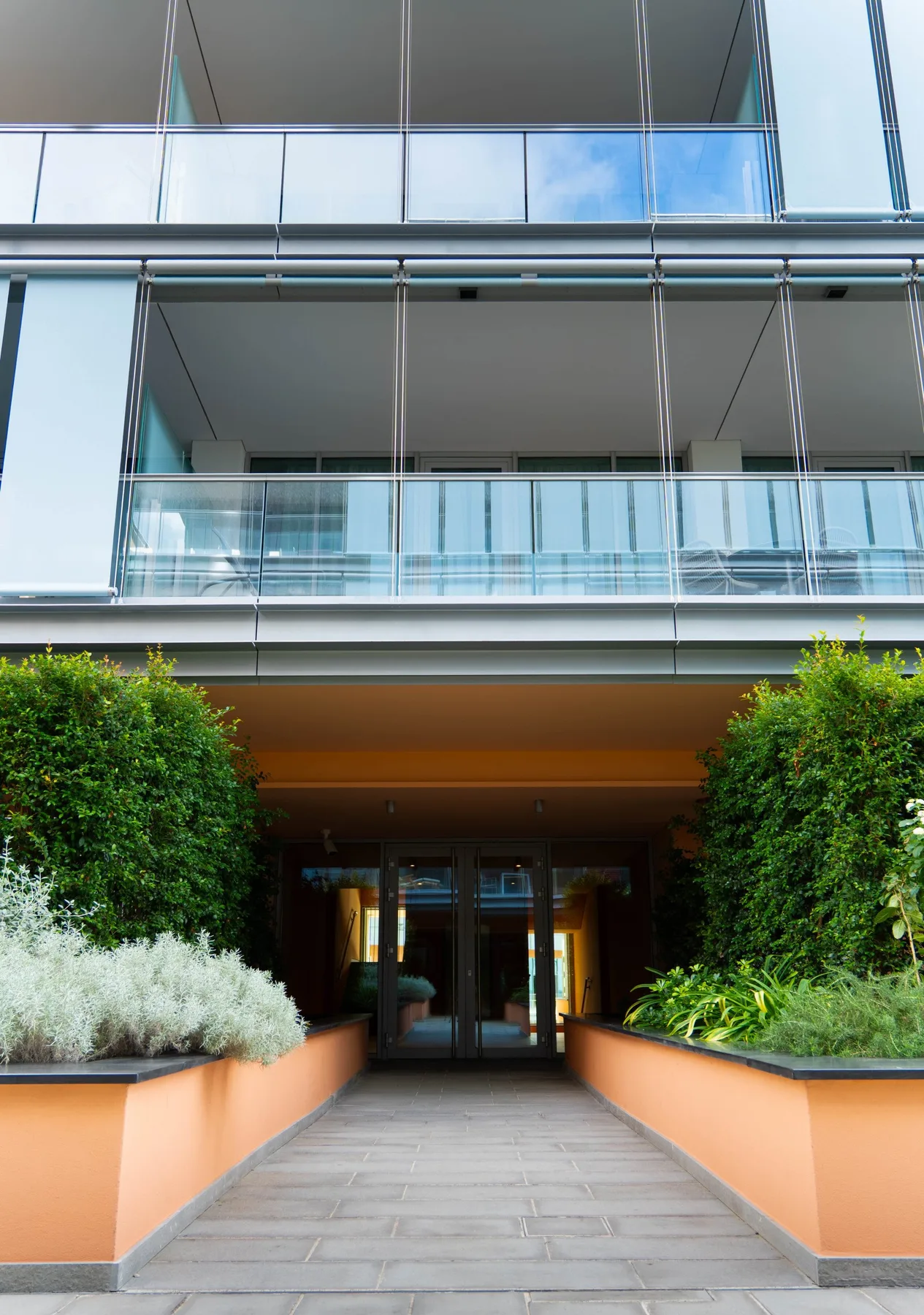 A building entrance is approached by a paved walkway bordered by planted beds.
