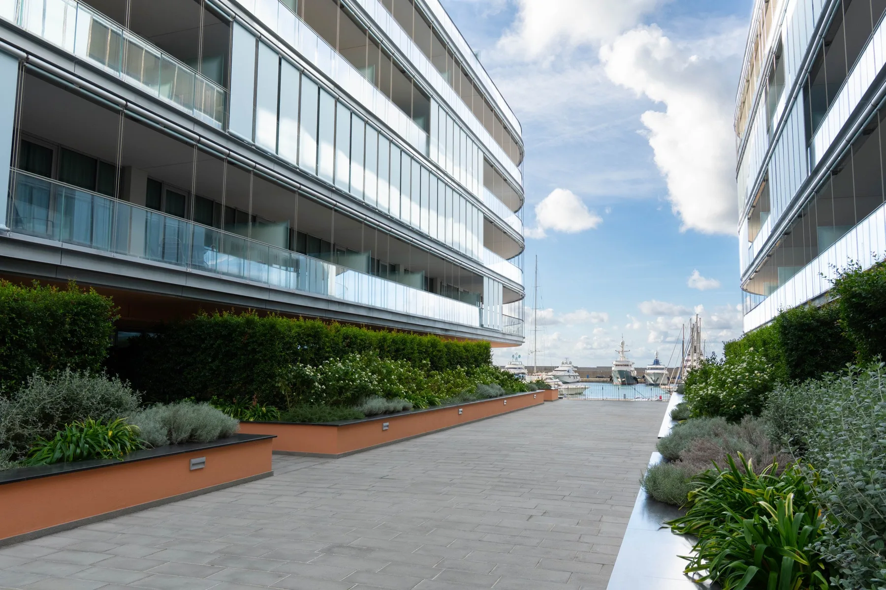 A paved walkway runs between modern buildings toward a waterfront.