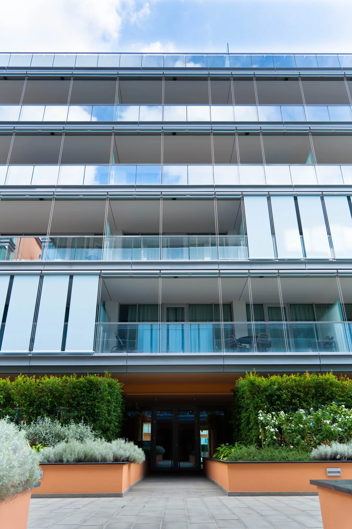A modern multistory building with glass balconies and landscaped planters at the entrance.