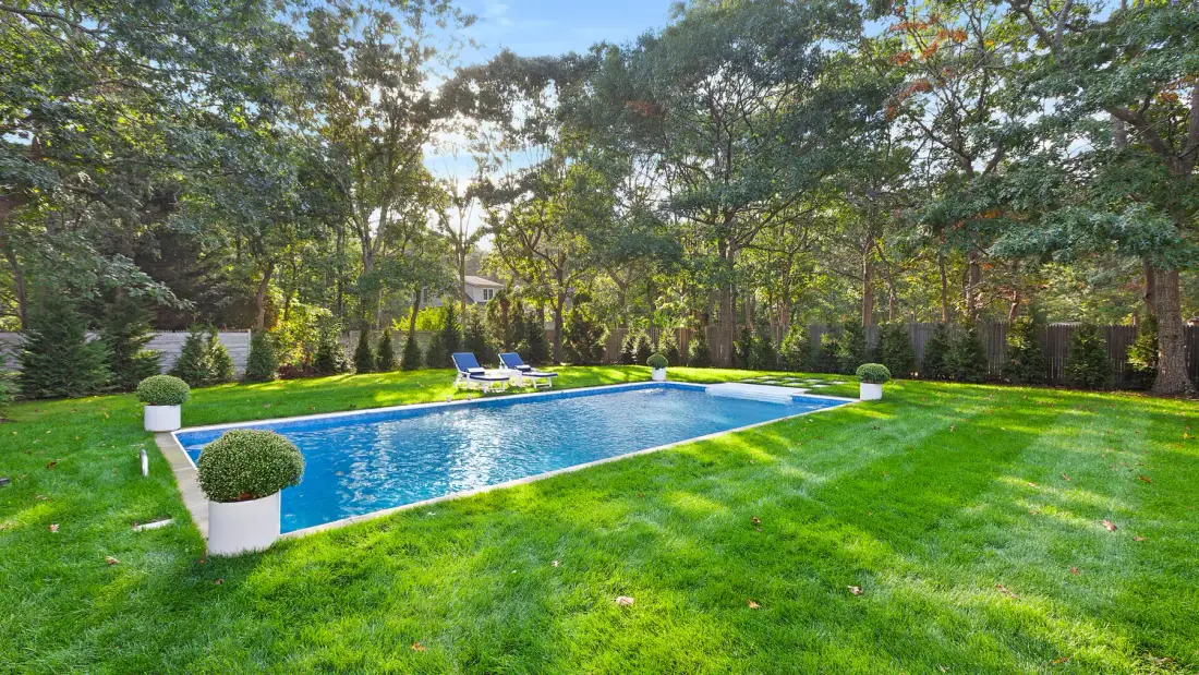 Una piscina rectangular está en un jardín con césped, rodeada de árboles.