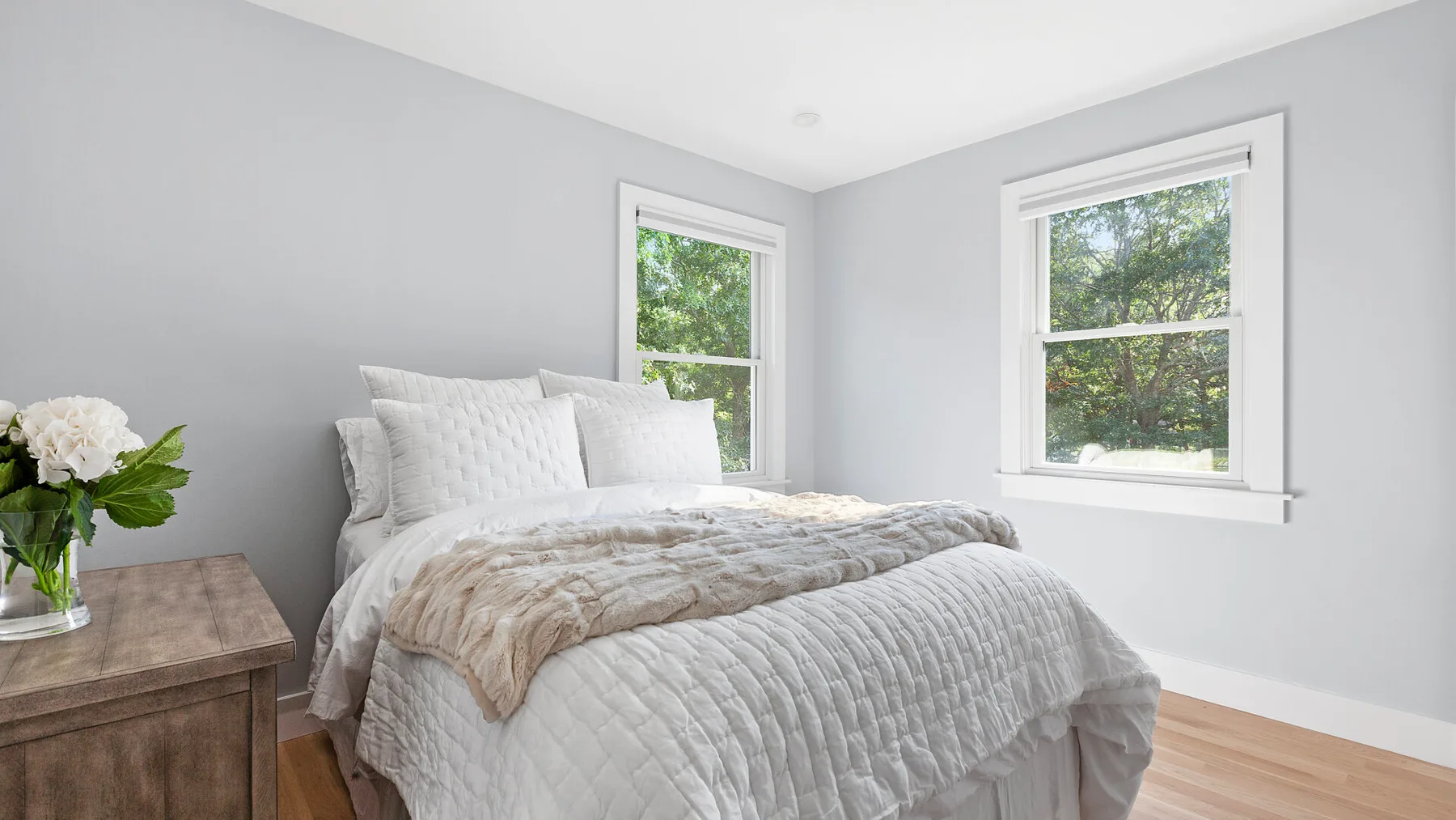 Un dormitorio luminoso con una cama vestida de blanco y dos ventanas.
