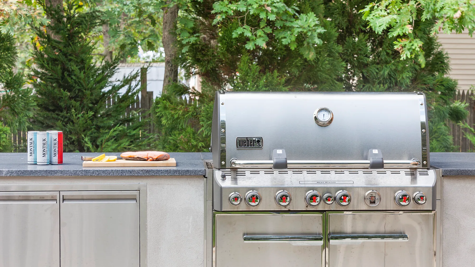 Una encimera de cocina exterior tiene una parrilla de gas de acero inoxidable bajo árboles frondosos.