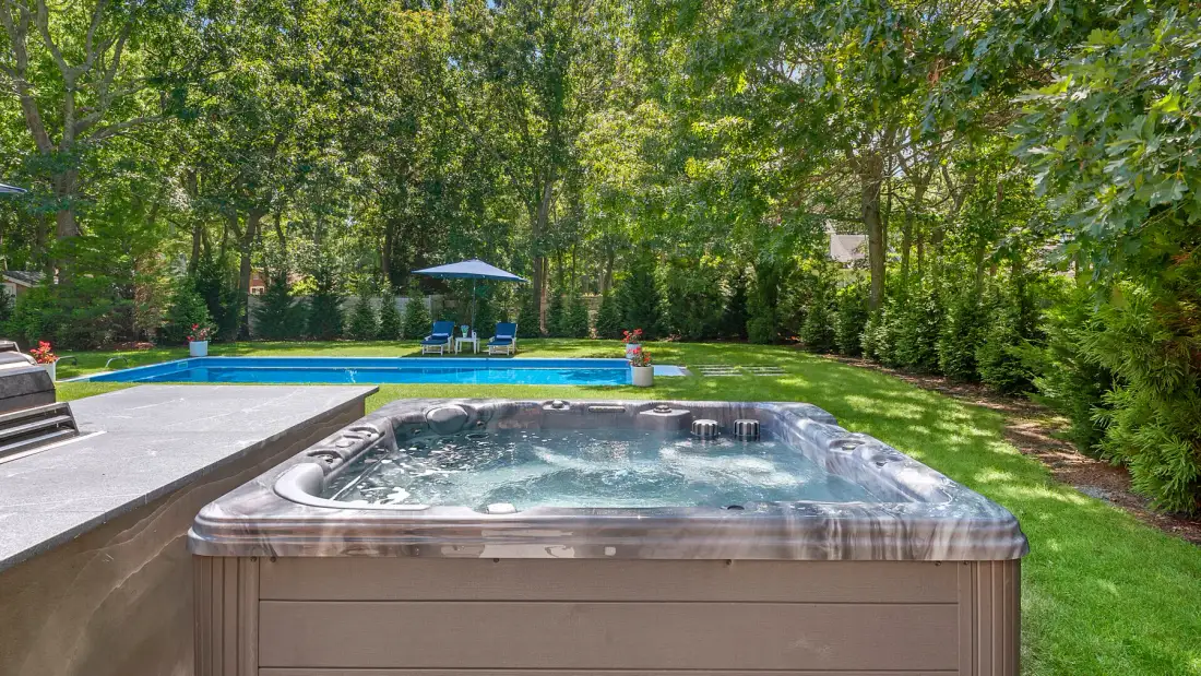Una bañera de hidromasaje exterior está junto a una piscina en un jardín frondoso.