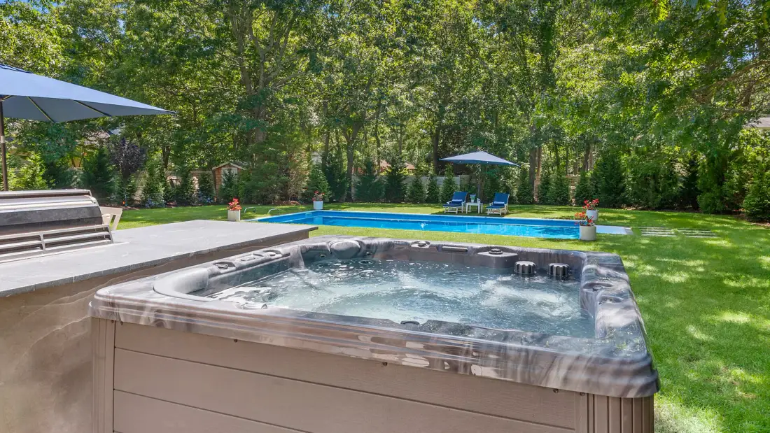 Una bañera de hidromasaje con burbujas está en un patio de piedra junto a una piscina y césped.