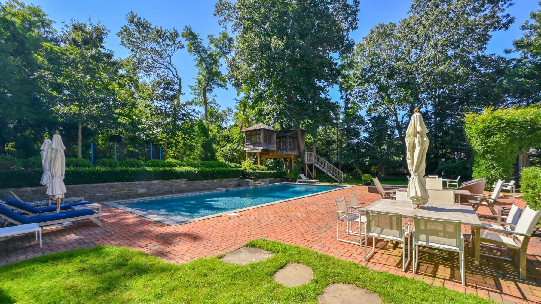 Una piscina al aire libre y un patio con muebles de comedor están rodeados de árboles.