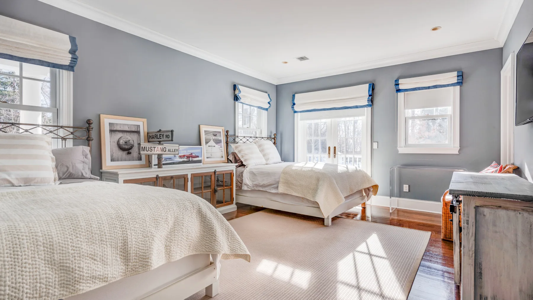 Un dormitorio luminoso con dos camas, cuadros enmarcados y ventanas con cortinas azules.