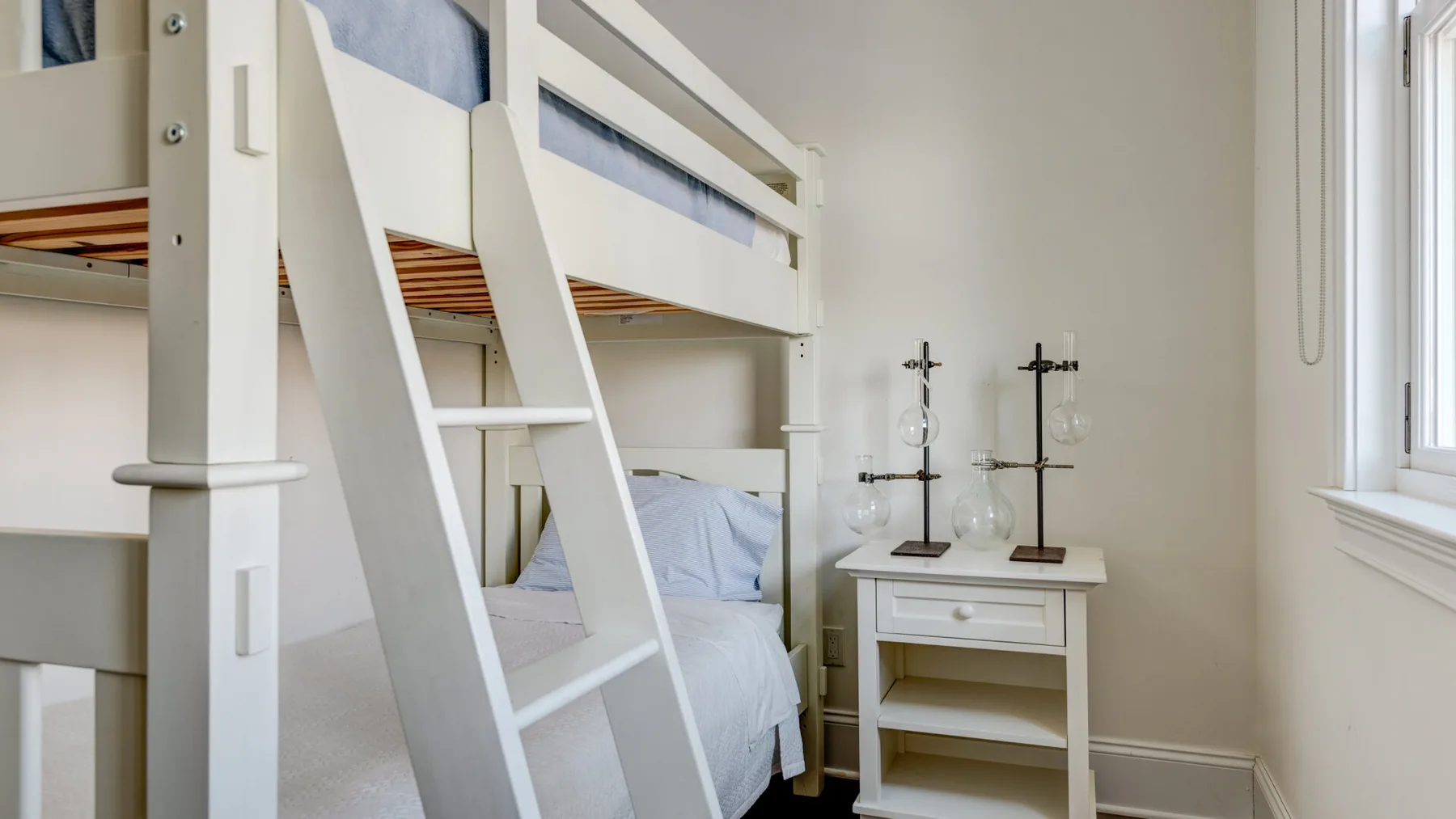 Un dormitorio luminoso con una litera blanca, una mesita de noche y una ventana.