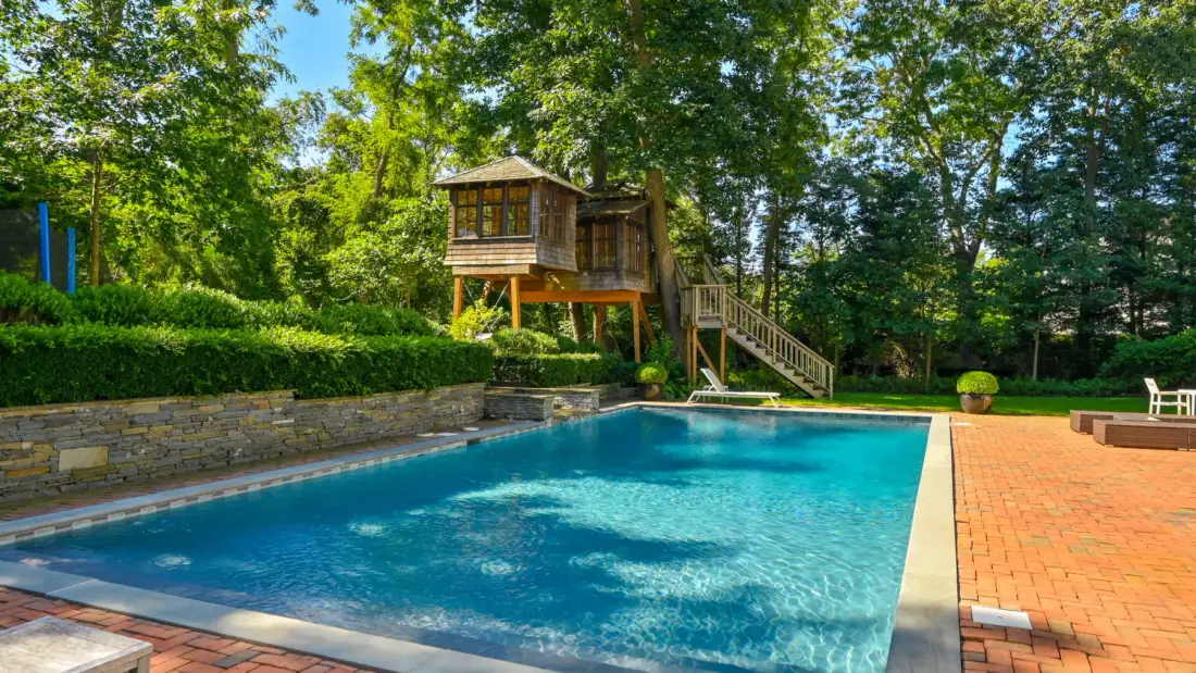 Una piscina al aire libre junto a un patio de ladrillo, con una casita de madera entre los árboles.