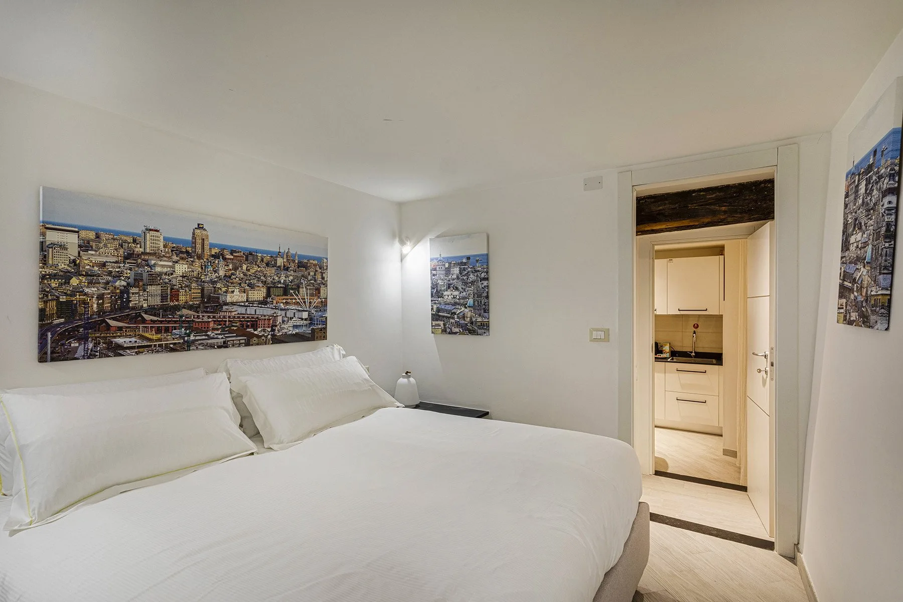 Un dormitorio luminoso con una cama blanca y cuadros de paisajes urbanos.