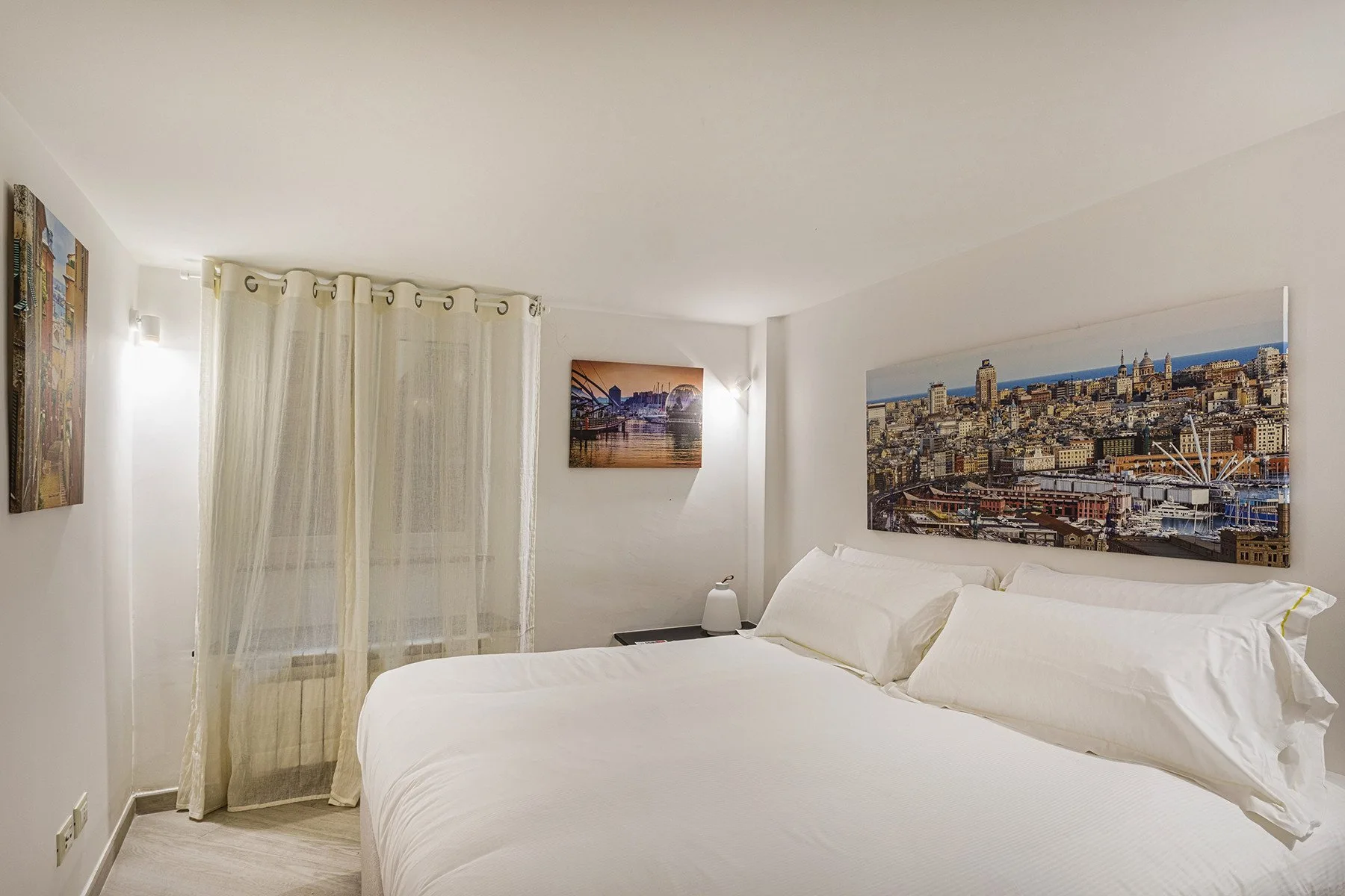 Un dormitorio con una cama bien tendida, cortinas translúcidas y una imagen urbana.