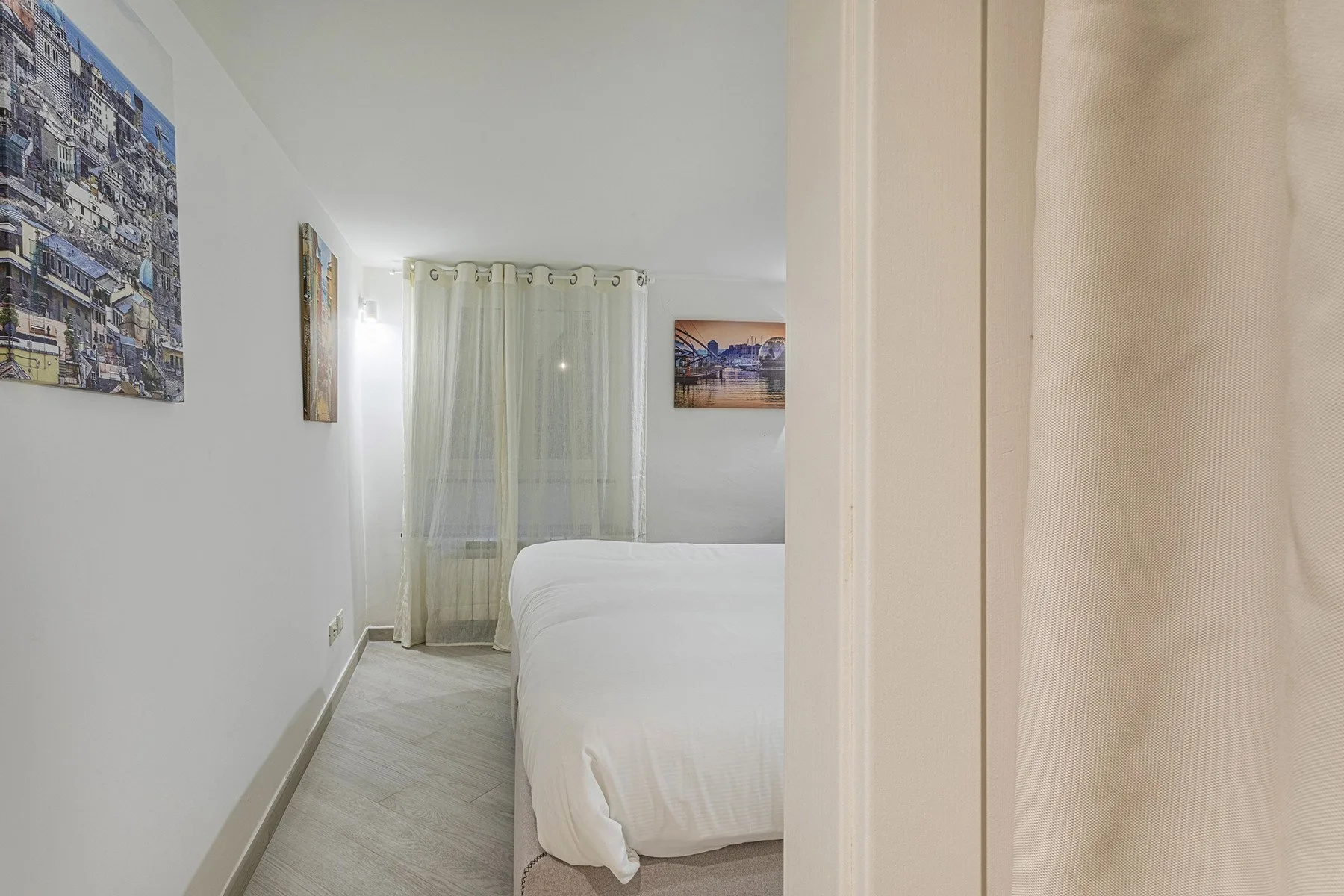 Un dormitorio con una cama, cortinas claras y cuadros en la pared.
