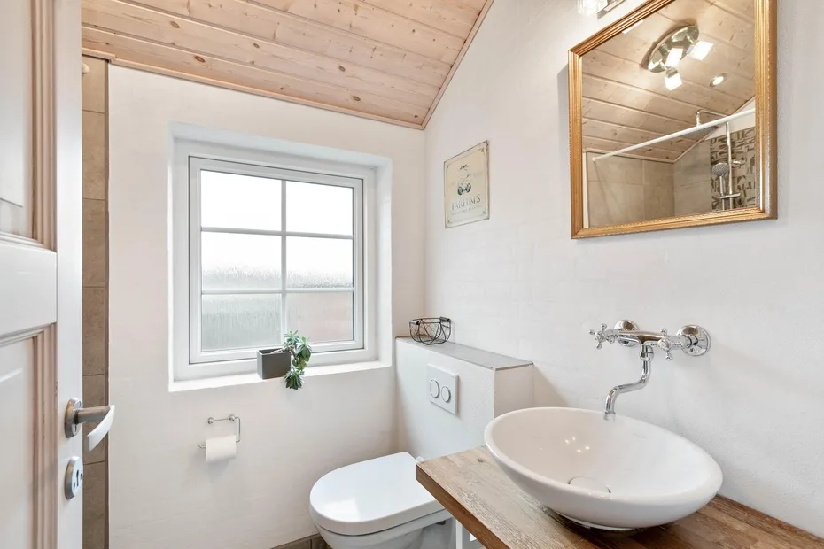 Un baño luminoso con ventana, inodoro y lavabo bajo un techo revestido de madera.